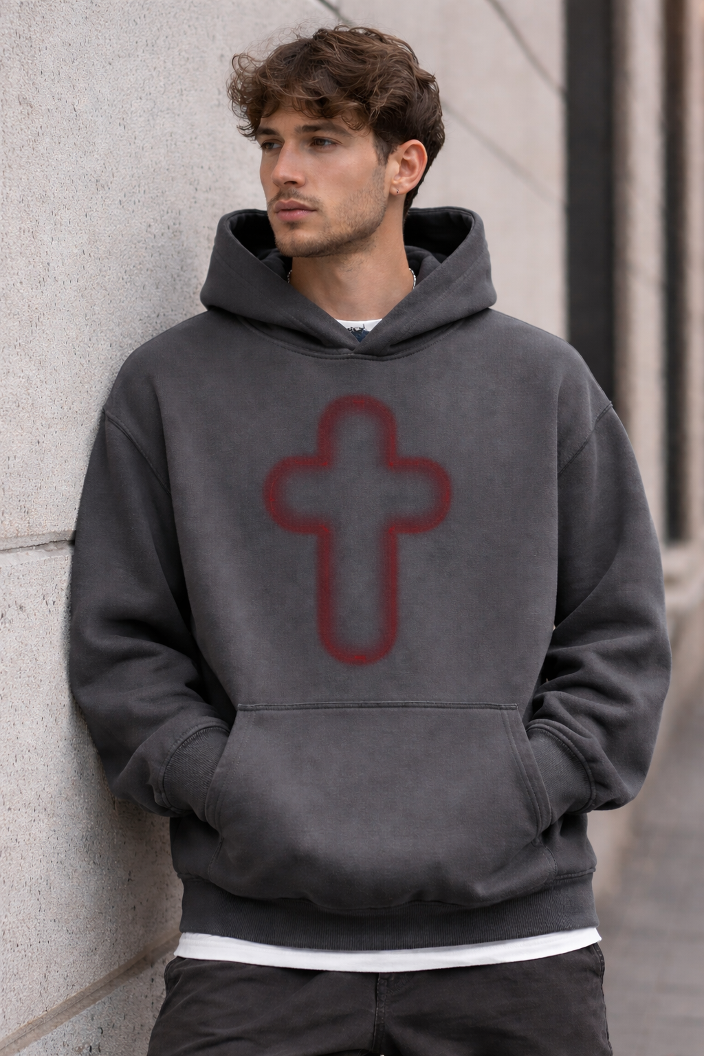 A young man with curly brown hair and blue eyes, wearing a dark gray hoodie with a red cross symbol on the front, standing outdoors against a beige wall.