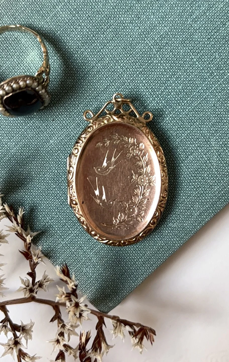 An antique oval pocket watch with a floral engraved design on its copper face, placed on a teal fabric background, with a branch of small dried white flowers nearby.