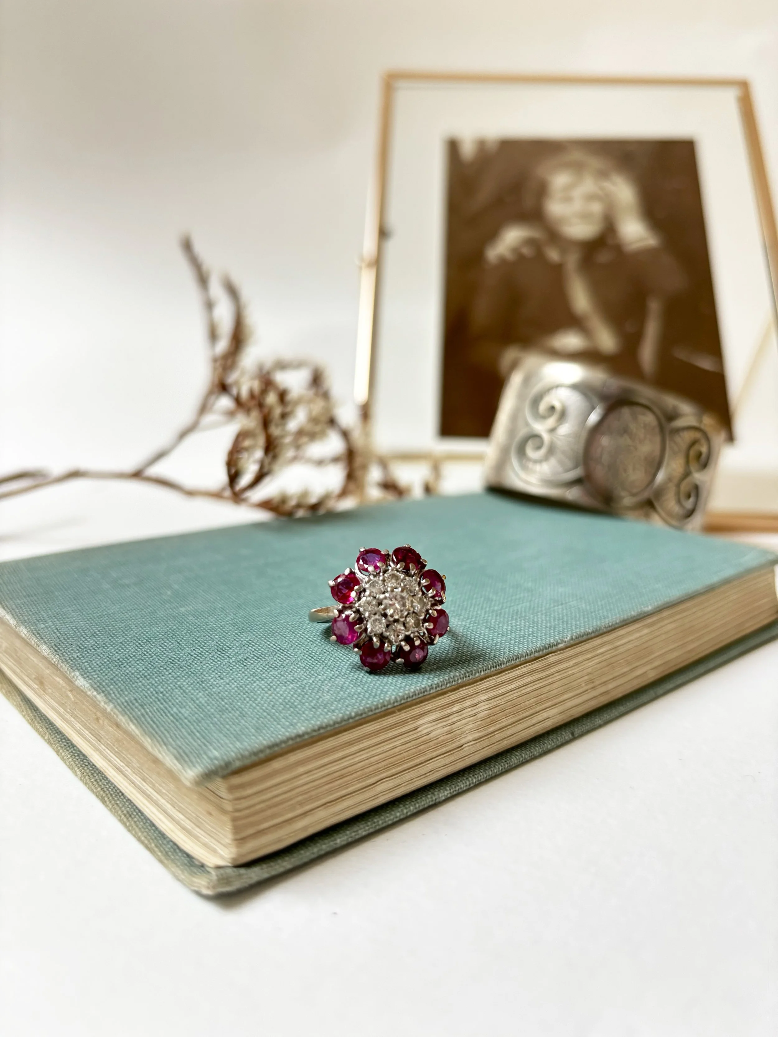 Close-up of a silver and ruby ring on a closed book with a mint cover, with a blurred framed black-and-white photograph and a decorative metal object in the background.