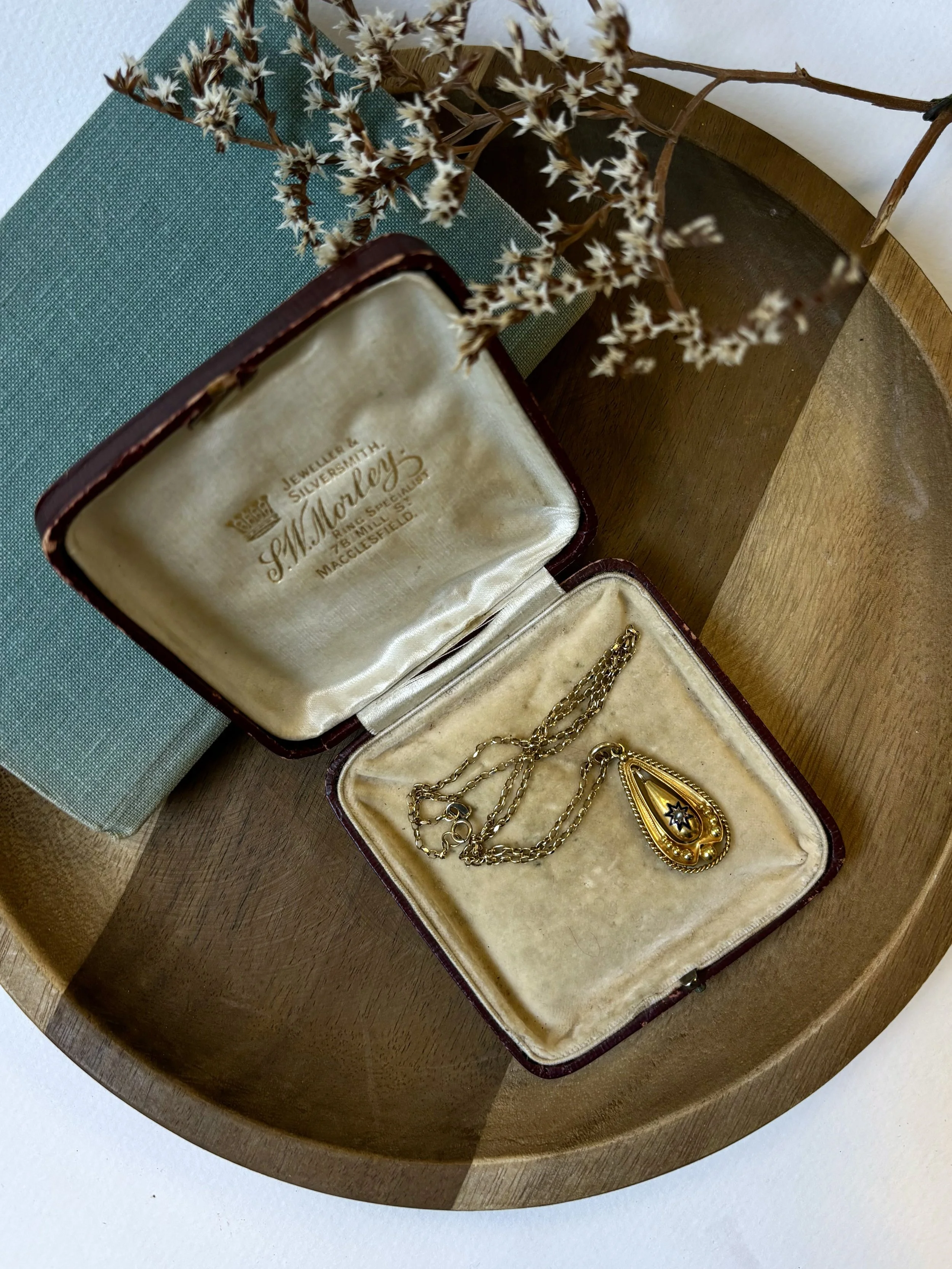 An open vintage jewelry box with a gold chain and a large teardrop-shaped gold pendant inside, placed on a wooden surface with a teal and a beige cloth nearby, and a bouquet of dried white and brown flowers.