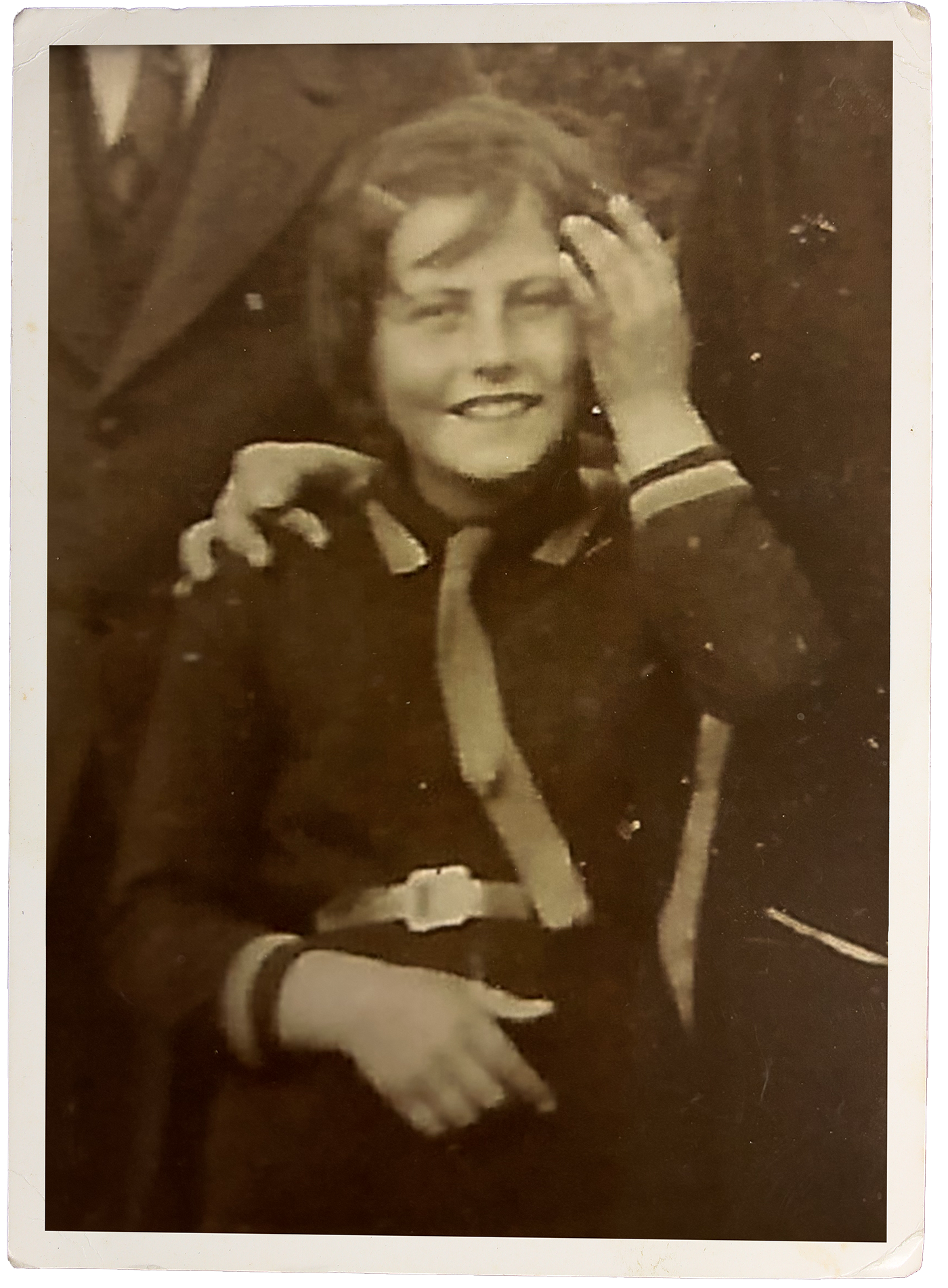 A sepia-toned vintage photograph of a smiling woman with short hair, wearing a sailor-style top with a stripe on the collar, and playing with her hair while sitting.