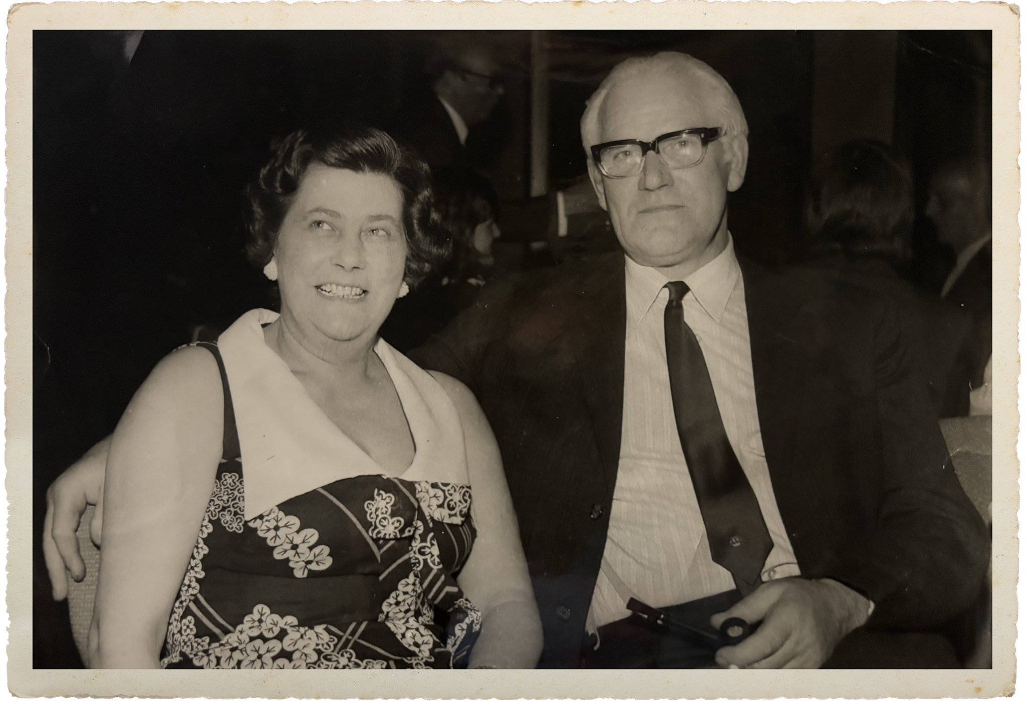 A black-and-white photograph of a smiling woman with short, styled hair and a man with glasses and white hair, both dressed in formal attire, at a social gathering.