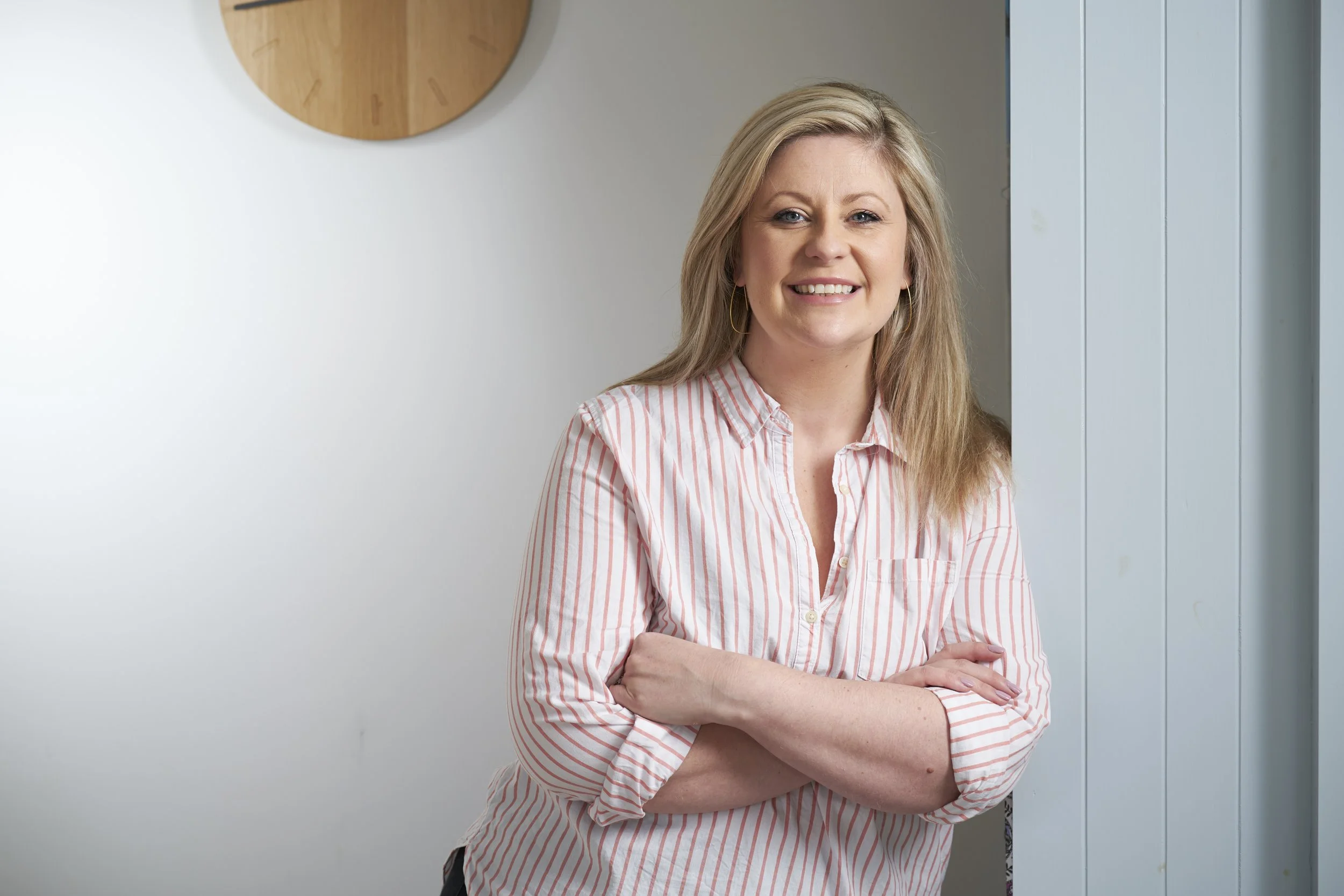 A woman with blonde hair wearing a pink and white striped shirt, smiling with arms crossed, standing in front of a white wall with a round wooden clock and a blue door.
