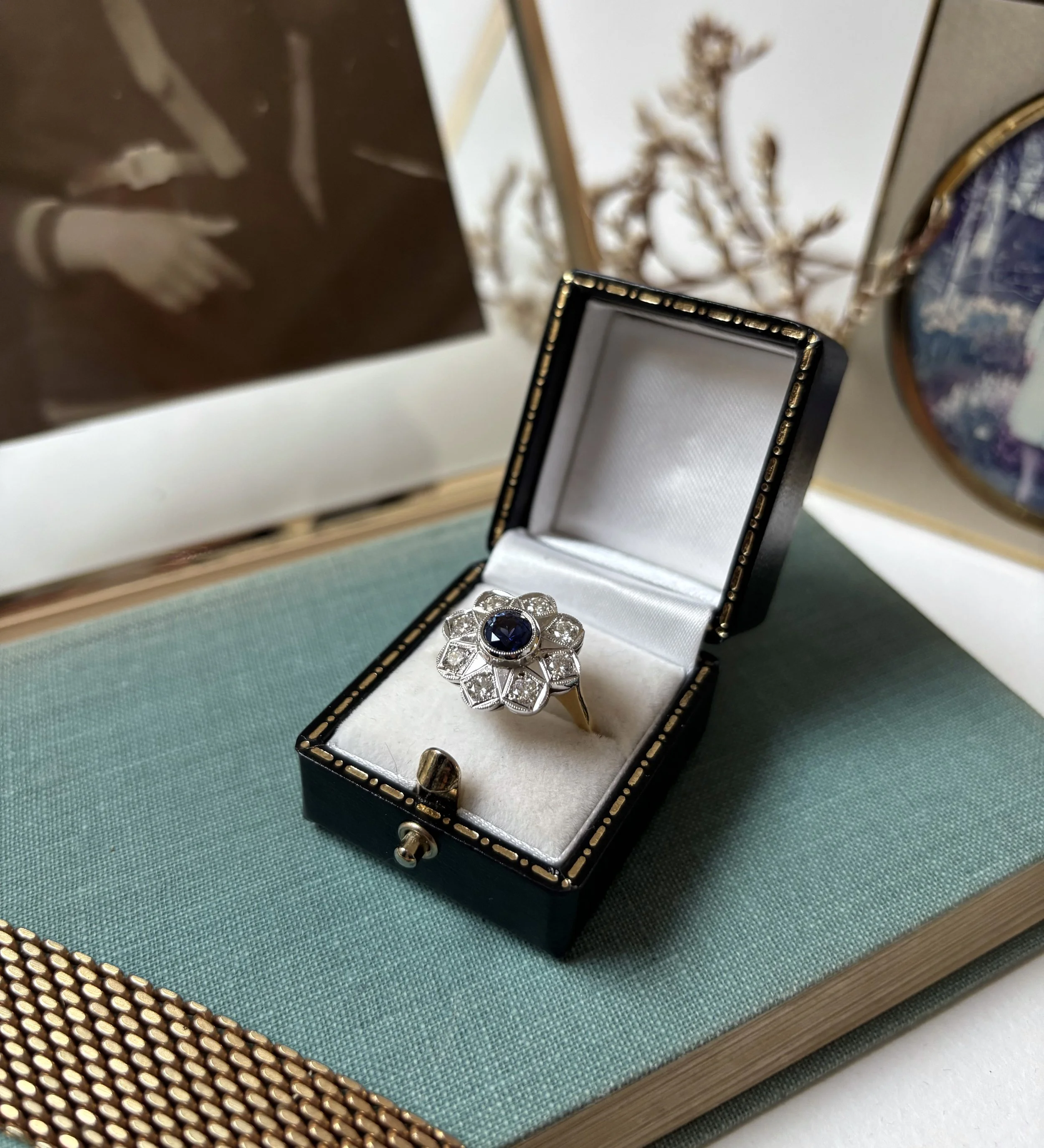 An open black jewelry box with a blue gemstone ring inside, positioned on a green fabric surface. Additional decor includes a brown box with a white silhouette of a woman in a trench coat, dried flowers in the background, and a decorative vase with a