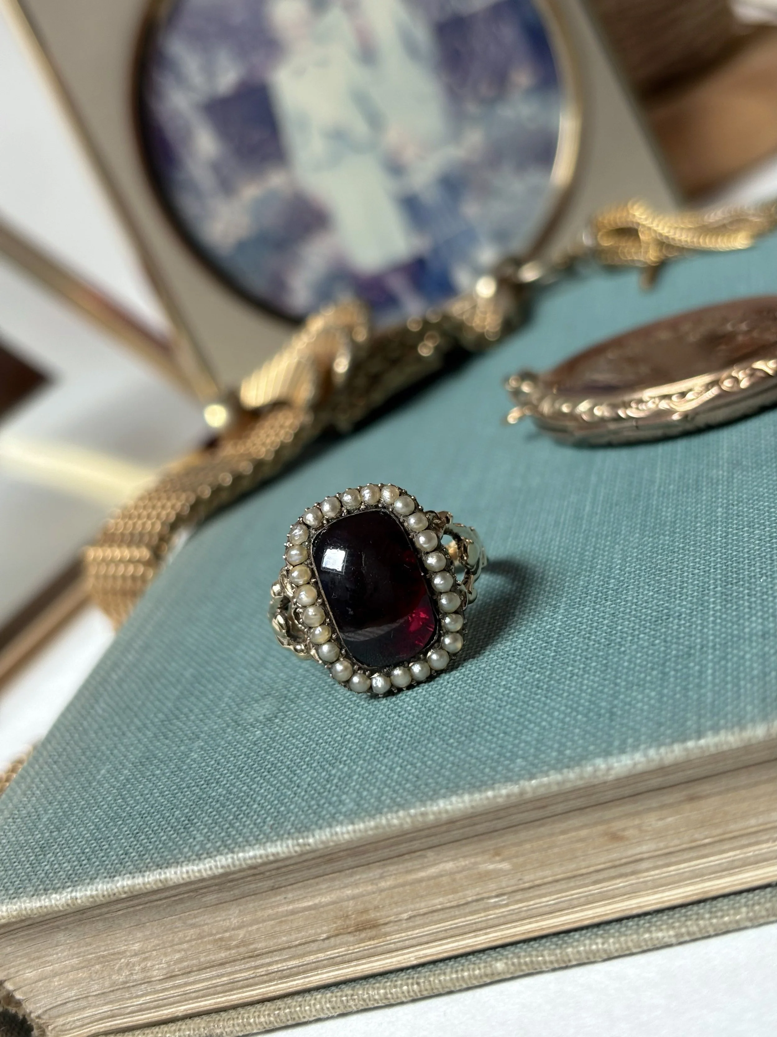 Close-up of vintage jewelry including a brooch with a red gem surrounded by pearls, a gold chain bracelet, and other jewelry pieces on a teal fabric surface with a book underneath.