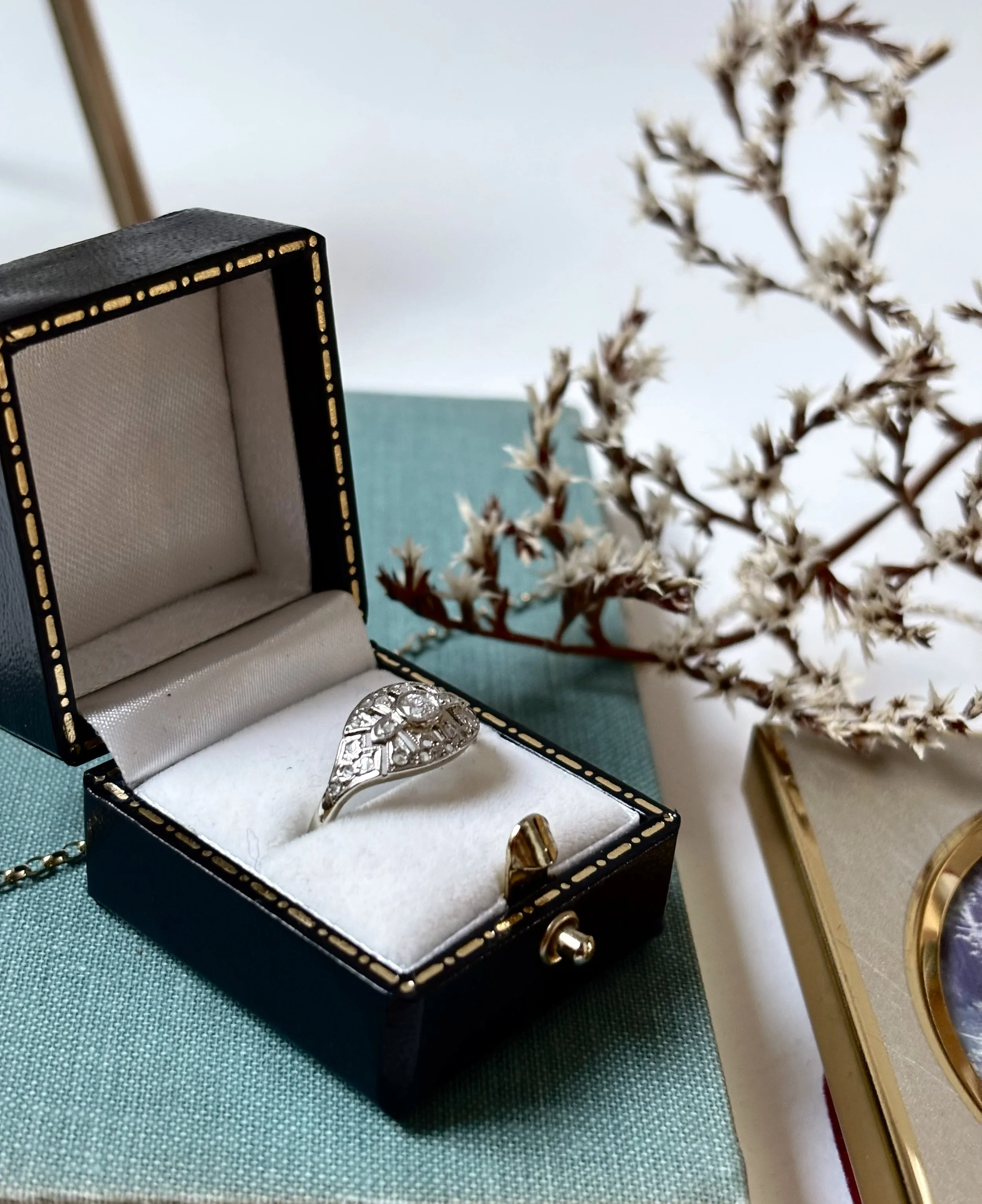 Open jewelry box containing a ring with a large stone, positioned on a green fabric surface. There is a decorative plant and a gold-colored object in the background.