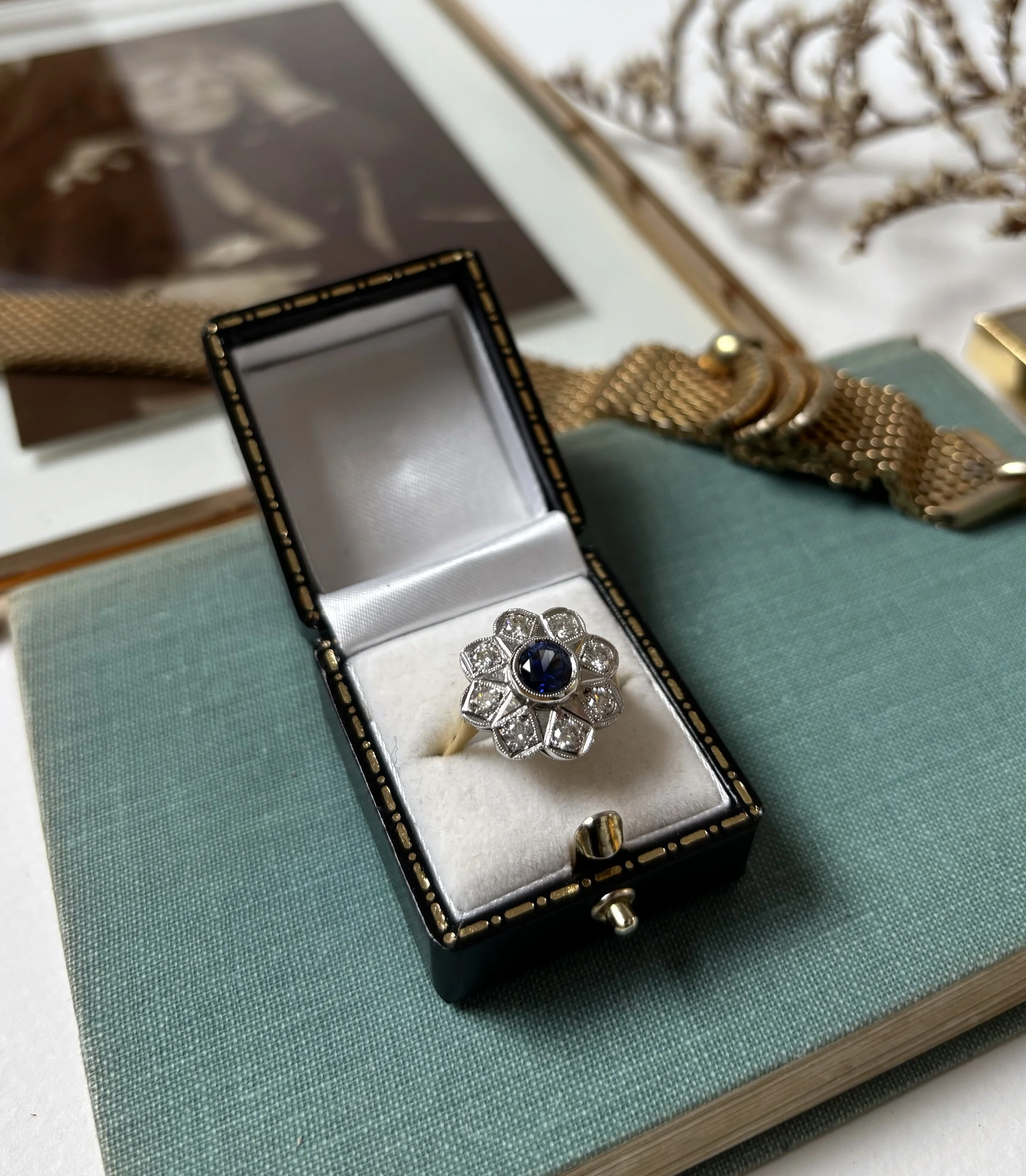 A jewelry box with an ornate flower-shaped ring featuring a large dark blue gemstone in the center, open on a fabric-covered surface with a dictionary and decorative items in the background.