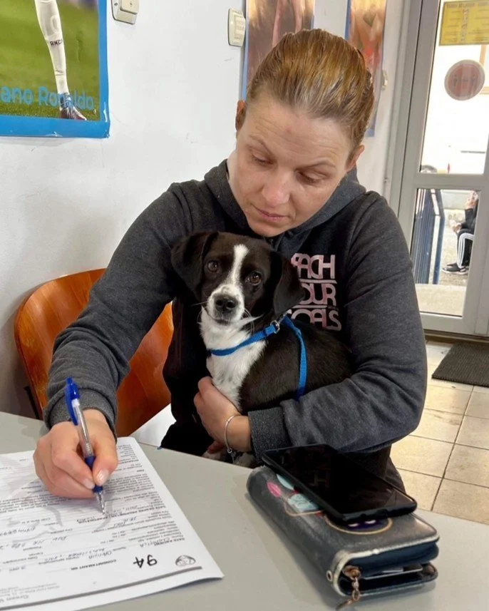 A woman sits at a table writing on a document. She has her arm around a small black dog sitting in her lap.