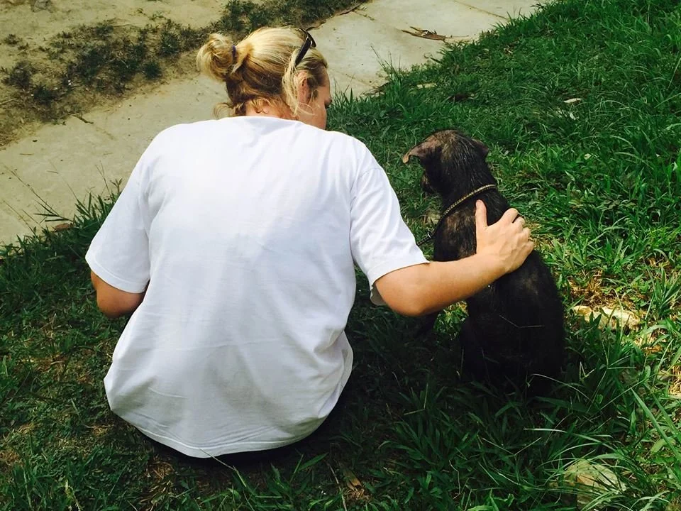 A woman sits on the grass with her arm around a black dog