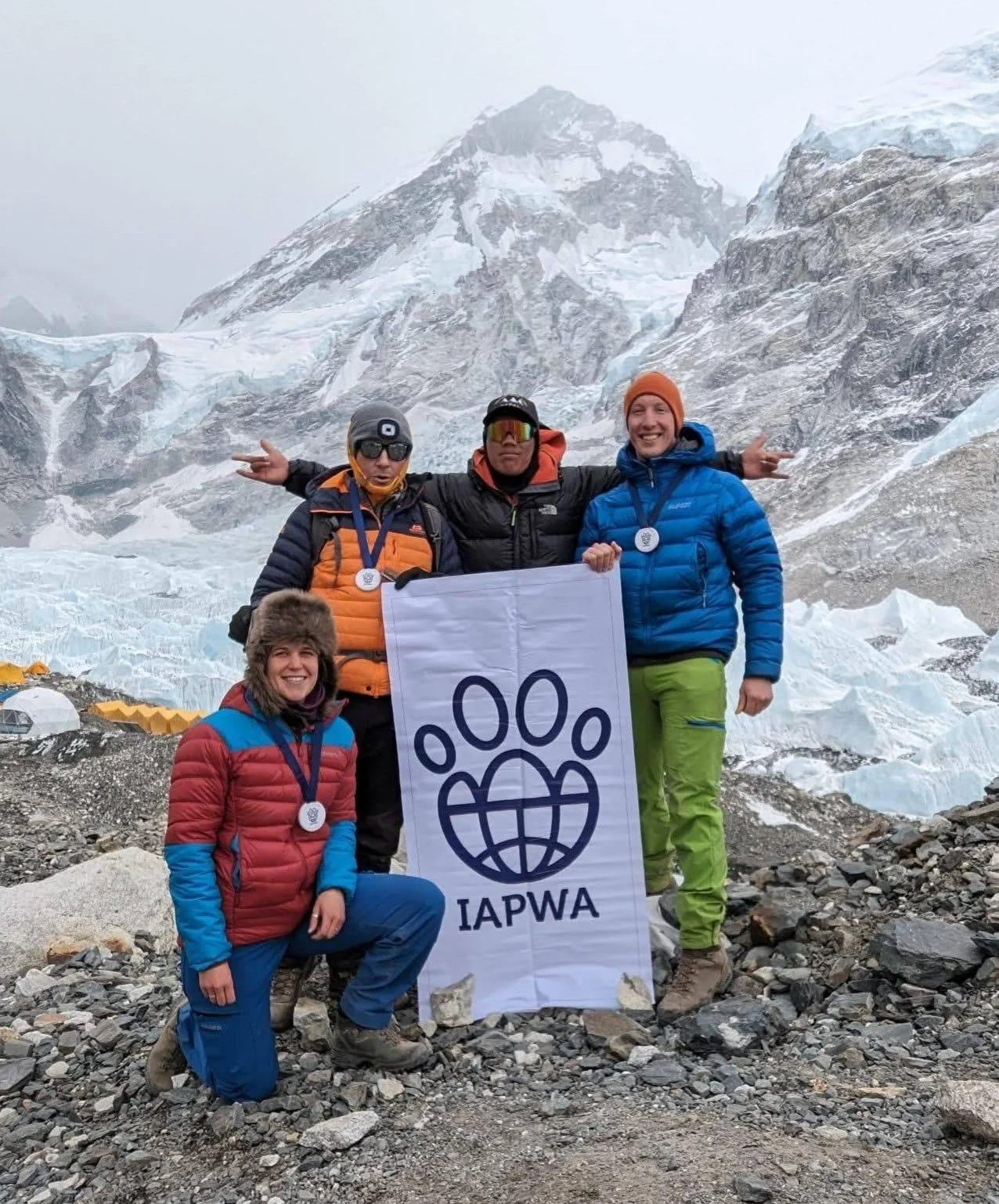Group of four people in outdoor gear against a stunning backdrop of snowy mountains, holding a flag with the IAPWA logo
