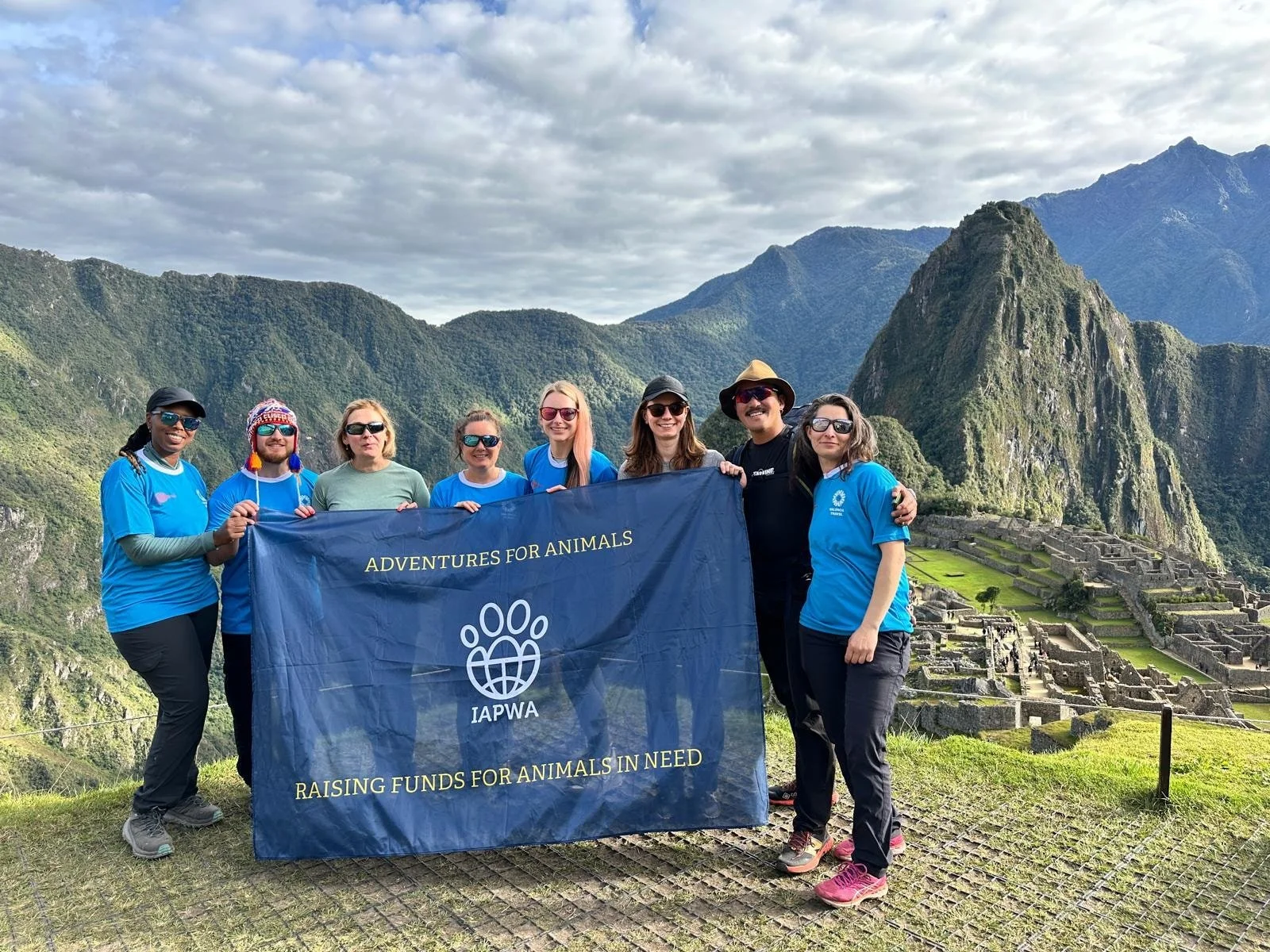 Group of people holding a blue banner with the words 'Adventures for Animals' and 'Raising funds for animals in need', standing outdoors in front of Machu Picchu in Peru.