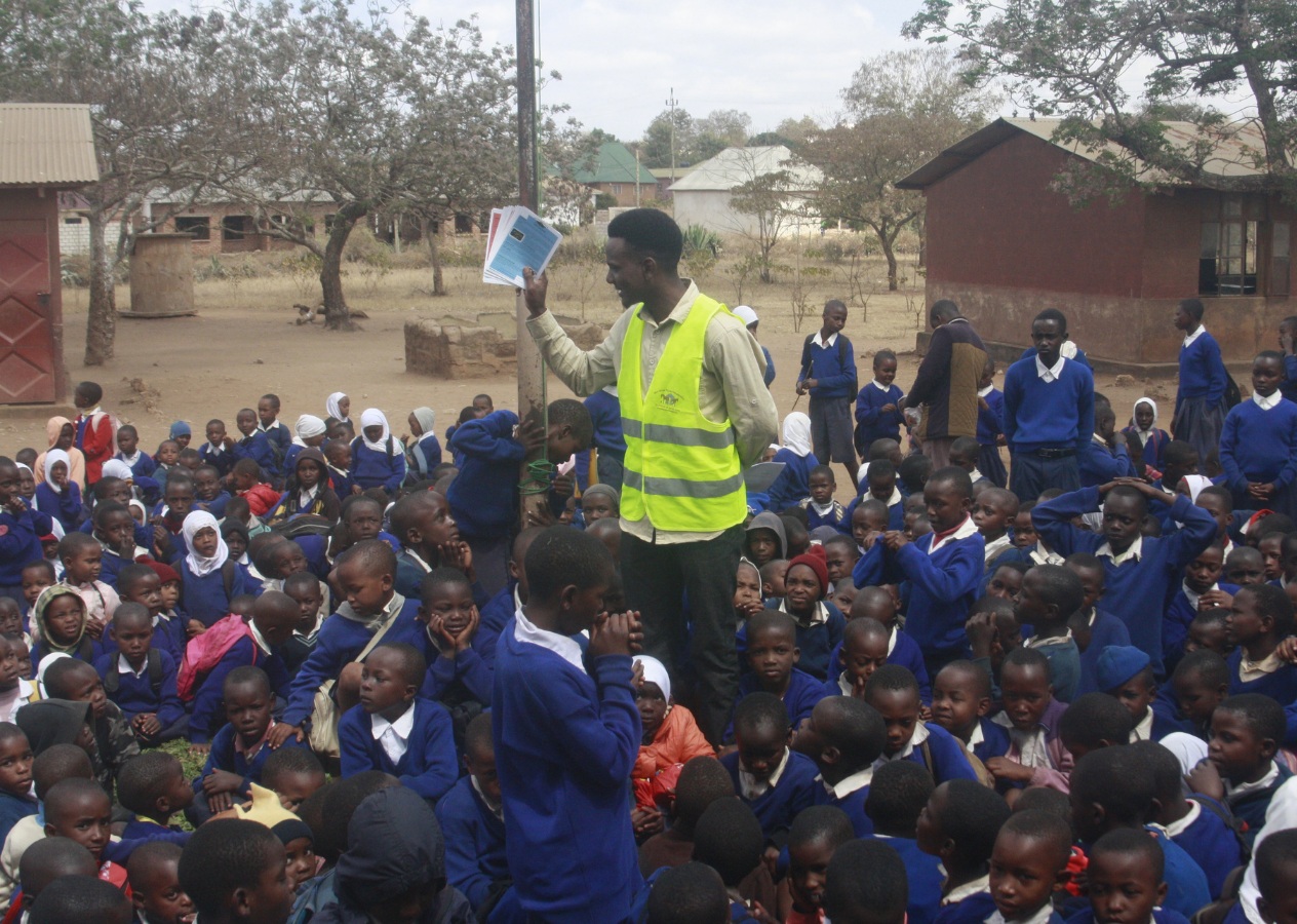 A man in a yellow safety vest stands among a large group of school children in blue uniforms, some sitting and others standing outside in a rural area with trees and buildings in the background.