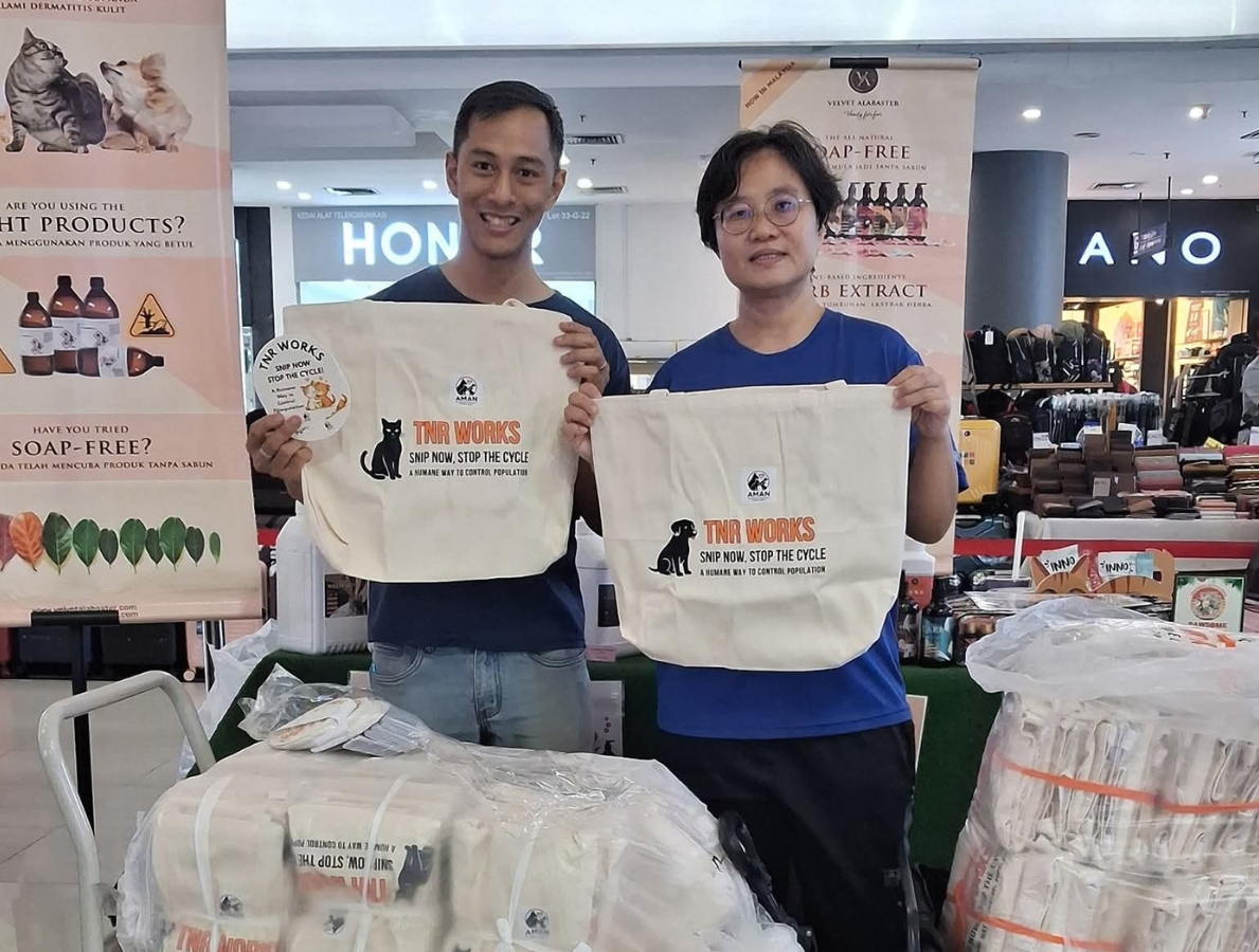 Two people standing behind a table holding tote bags with a logo that reads 'TNR WORKS. SNIP NOW, STOP THE CYCLE'