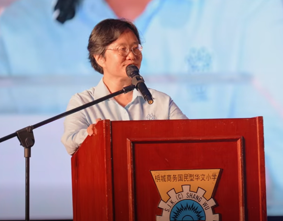 Woman speaking at a podium with a microphone, wearing glasses and a light-colored shirt, in front of a blue background.