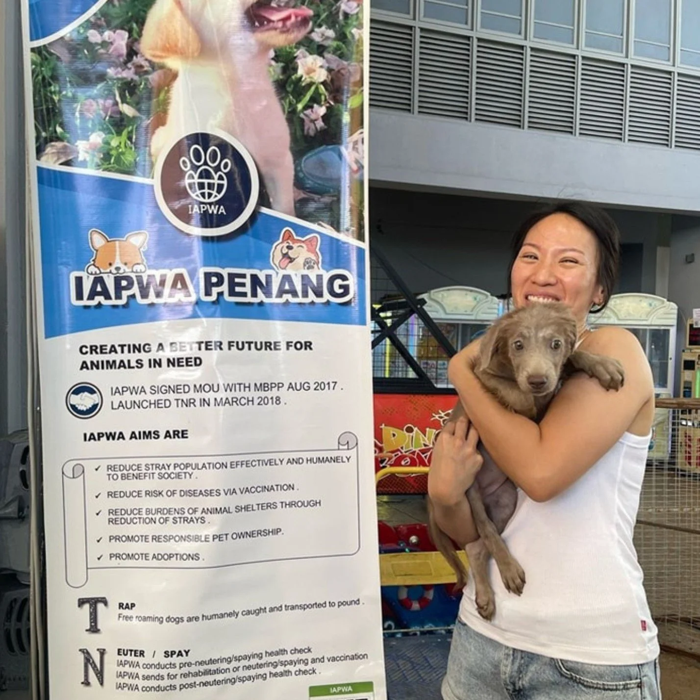 A woman smiling while holding a small puppy in front of a poster for IAPWA Penang