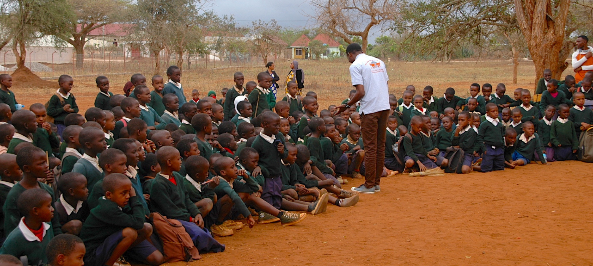 Group of schoolchildren sitting and kneeling on the ground outdoors, listening to a person speaking