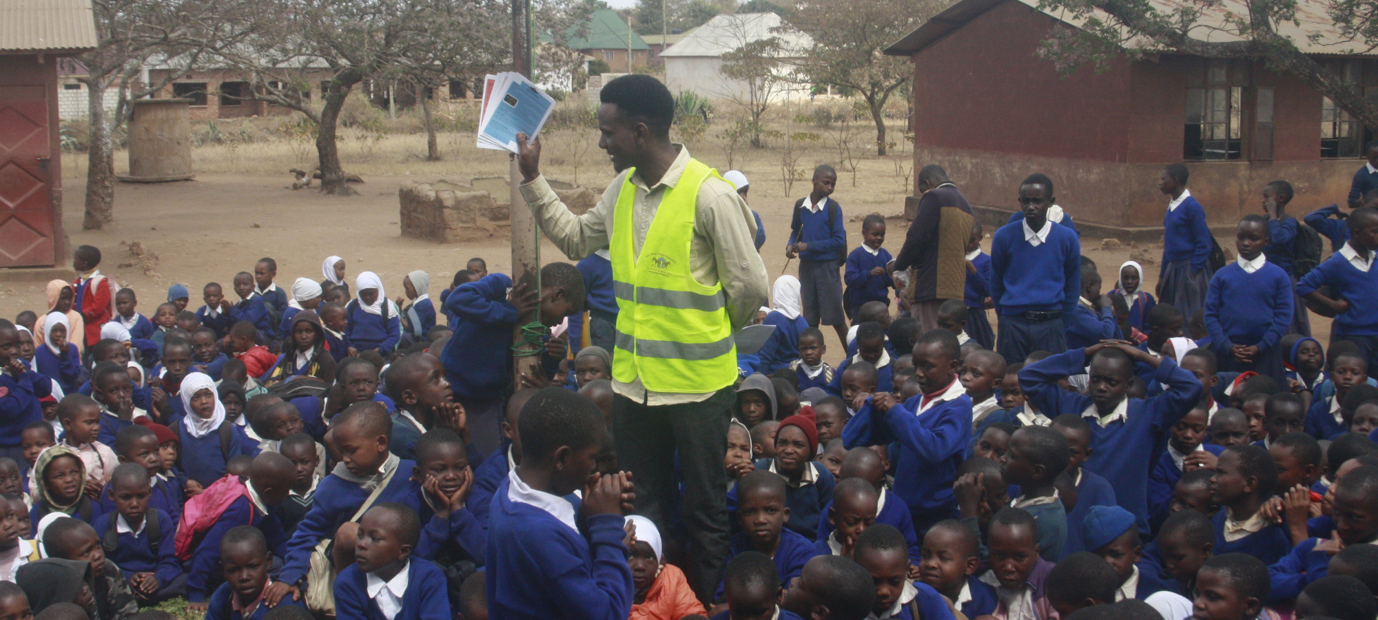 A man in a yellow safety vest holds up papers while standing among a large group of children in school uniforms outside.
