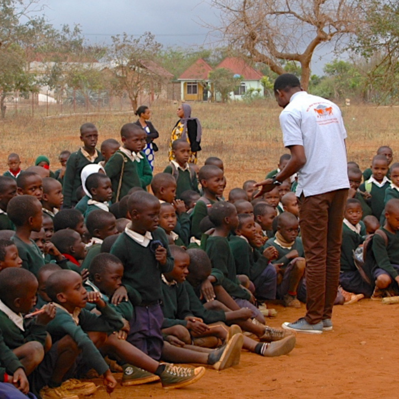 A group of school children in green uniforms sitting on the ground outdoors, with a man in a white shirt handing something to them, and two women standing behind, in a rural area with trees and small houses in the background.