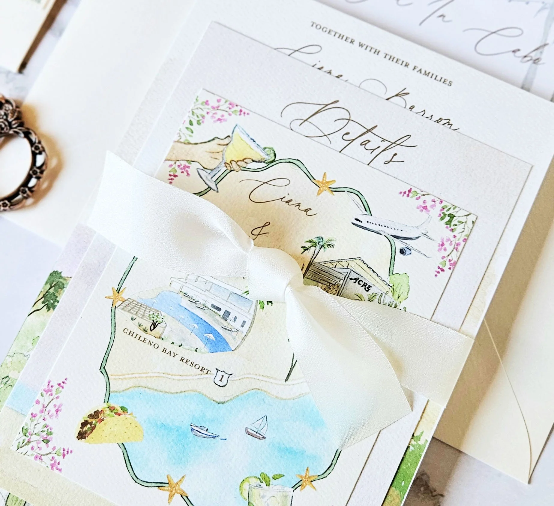 A wedding invitation with a colorful map illustration of Chileno Bay Resort, showing a pool, boats, and surrounding scenery, tied with a white ribbon.