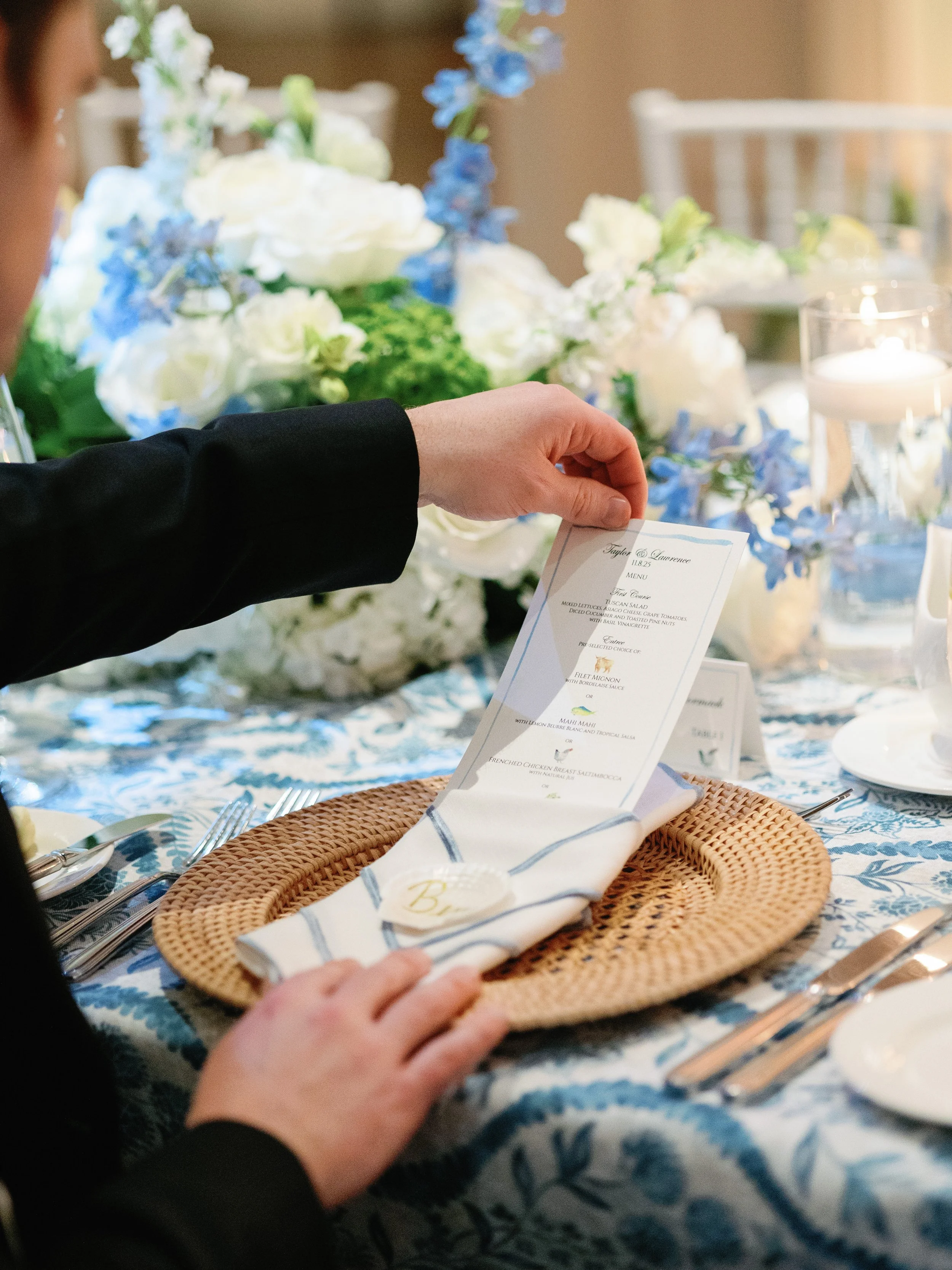 A person is holding a wedding menu card over a table setting with a woven placemat and a folded napkin, surrounded by floral arrangements and candles on a decorated table.
