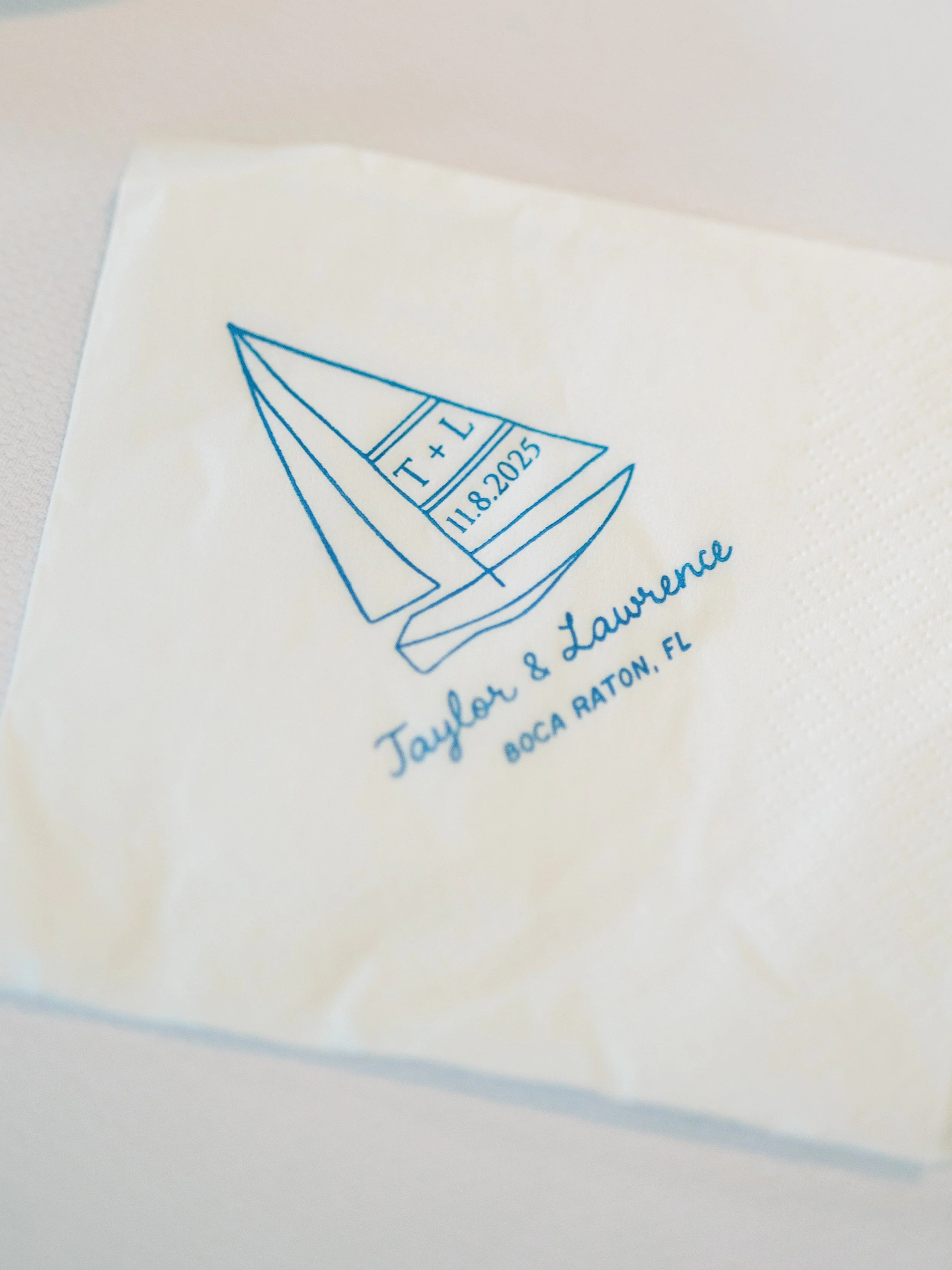 A napkin printed with a blue outline drawing of a sailboat with the date "11.8.2025" and initials "T+L" on the sail, along with the names "Taylor & Lawrence" and the location "Boca Raton, FL".