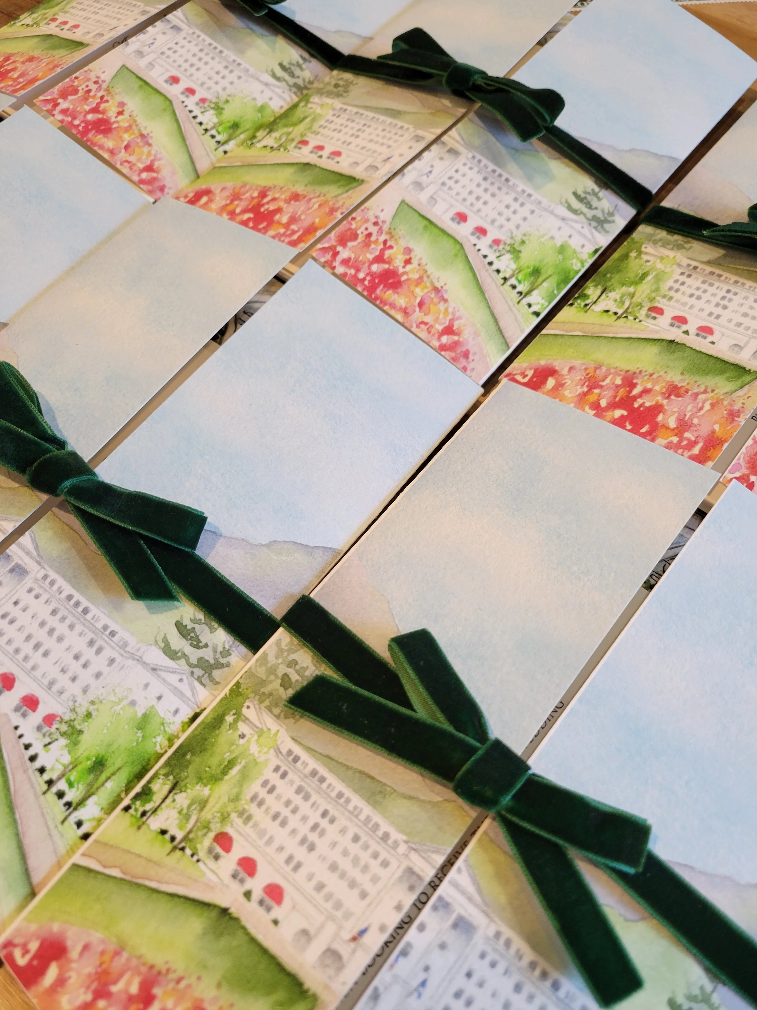 Sets of greeting cards or invitations with watercolor painting of a cityscape, greenery, and pink flowering trees, tied with green velvet ribbons.