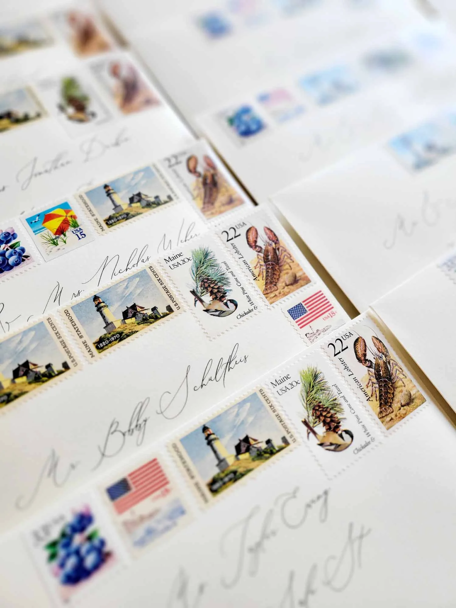 Close-up of envelopes with American flag stamps, lighthouse stamps, and lobster stamps, addressed with cursive handwriting.