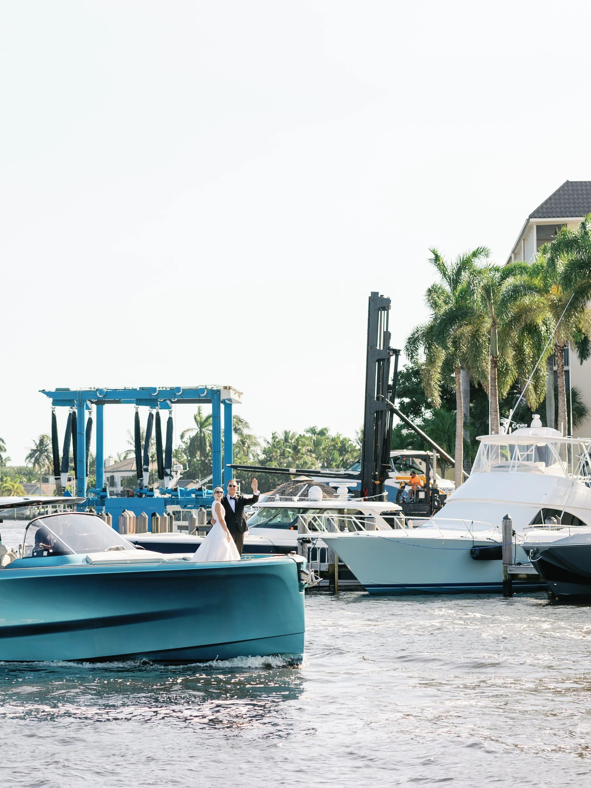 A wedding couple waving from a boat at a marina with yachts, palm trees, and a building in the background.