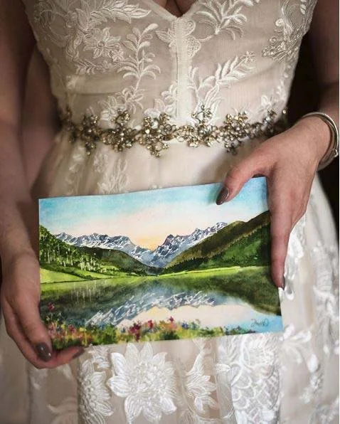 A woman in a white dress with floral embroidery and beaded belt is holding a painting of a mountain landscape with a lake and colorful flowers in the foreground.