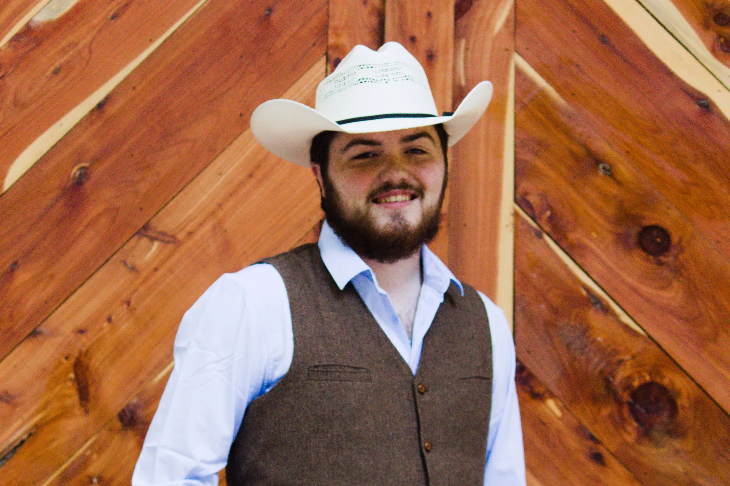 A man wearing a white cowboy hat, white shirt, and brown vest, smiling, standing in front of a wooden background.