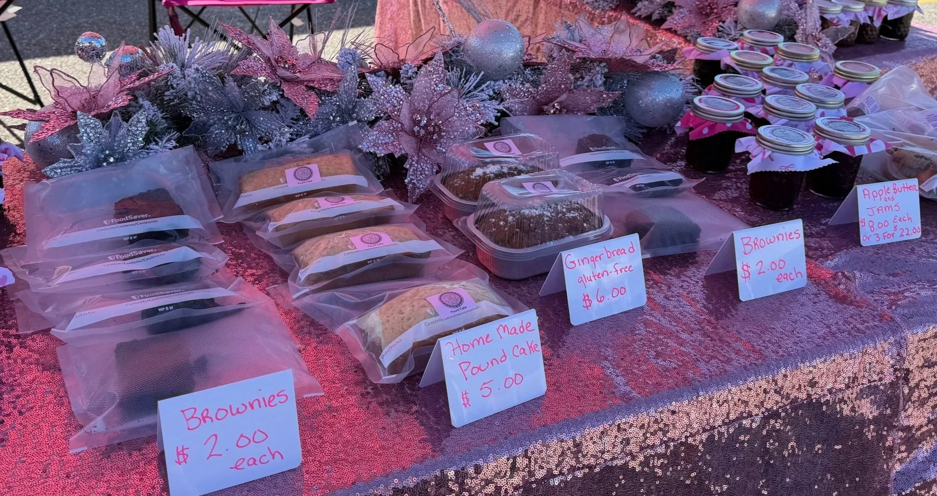 A display of homemade baked goods on a table decorated with pink and silver Christmas ornaments and plants. The baked goods include brownies, gingerbread, apple butter jam, and pound cake, each labeled with handwritten signs indicating prices.