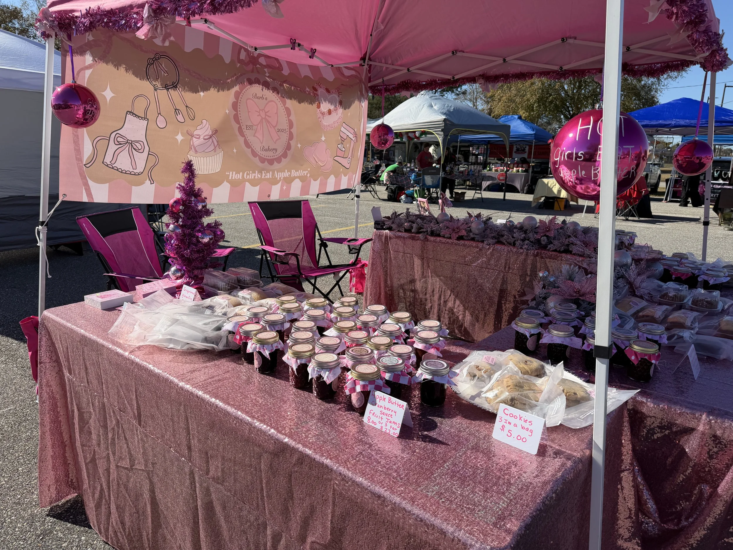 Market stall decorated in pink with jars of apple butter, cookies, and pink balloons, at an outdoor fair or market.