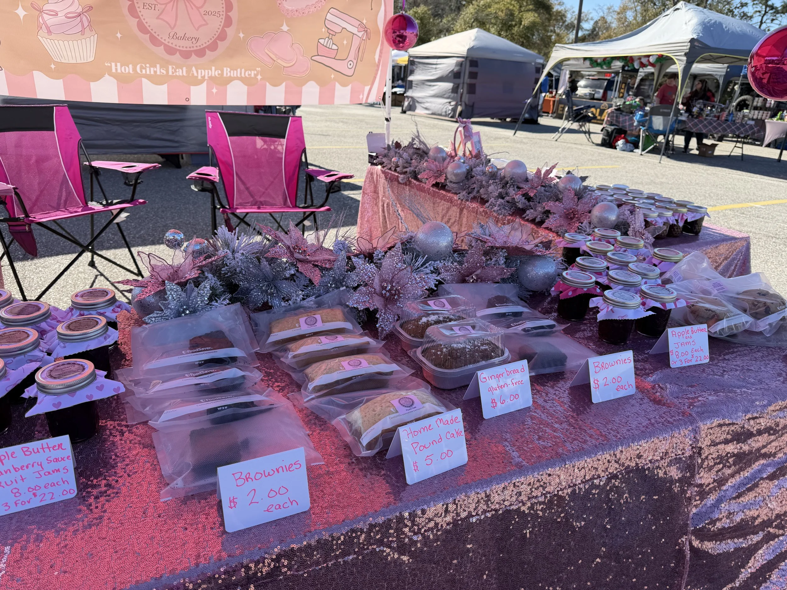 Outdoor market stall decorated with pink ornaments and festive foliage, displaying baked goods including brownies, gingerbread pound cake, apple butter and jams, with signs showing prices in dollars.