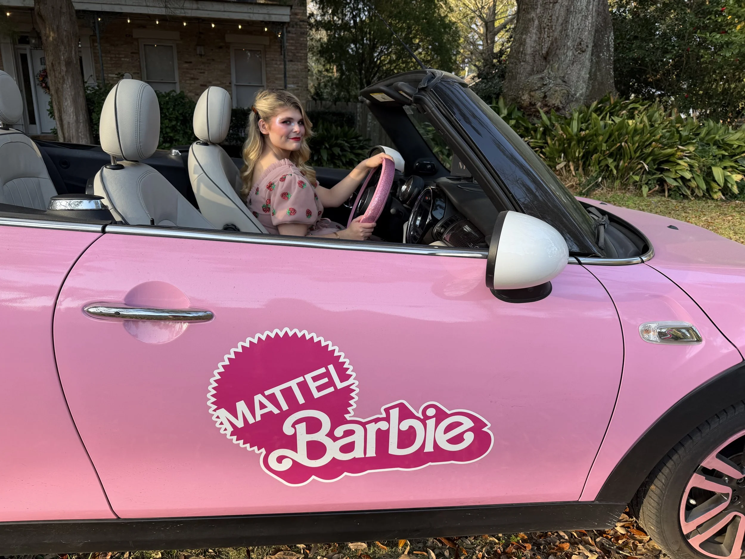 A young woman with blonde hair and a pink dress with strawberries, sitting in the driver's seat of a pink convertible car with a Mattel Barbie logo on the door, parked outdoors with greenery and a house in the background.