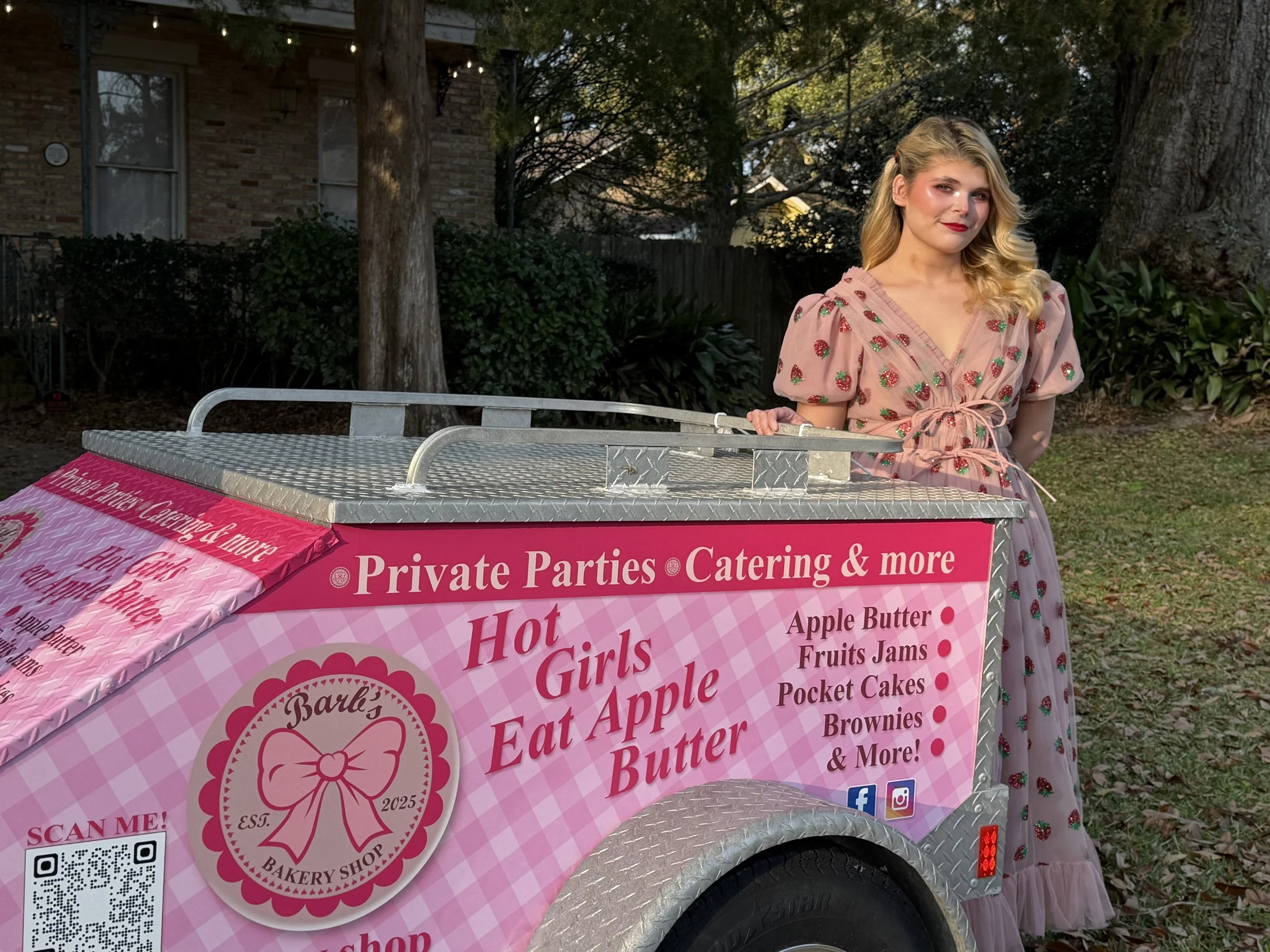 A woman with long blonde hair and pink lipstick stands next to a pink and silver bakery cart with a sign that reads 'Hot Girls Eat Apple Butter.' She is wearing a pink dress with a strawberry pattern. Trees and a house are visible in the background.