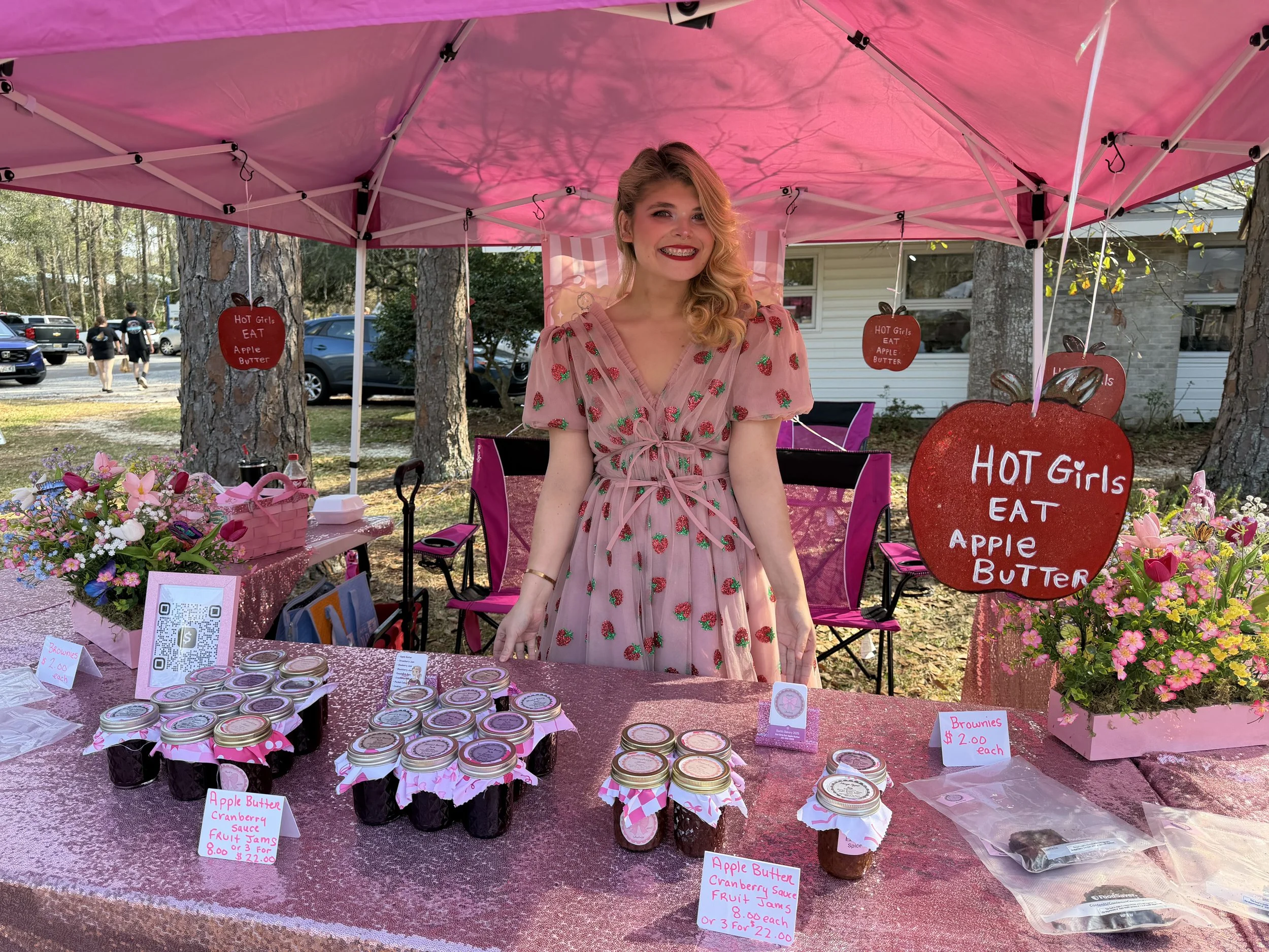 Woman standing behind a table at a pink-themed outdoor market stall with jars of apple butter and signs that say "HOT Girls EAT APPLE BUTTER" and "Browines $2.00 each." She is wearing a pink dress with strawberry patterns and smiling.