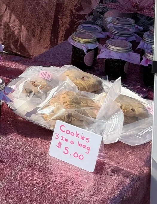 Cookies in clear plastic bags on a table, with a sign that reads 'Cookies 3 in a bag $5.00'; jars of jam or jelly in the background.