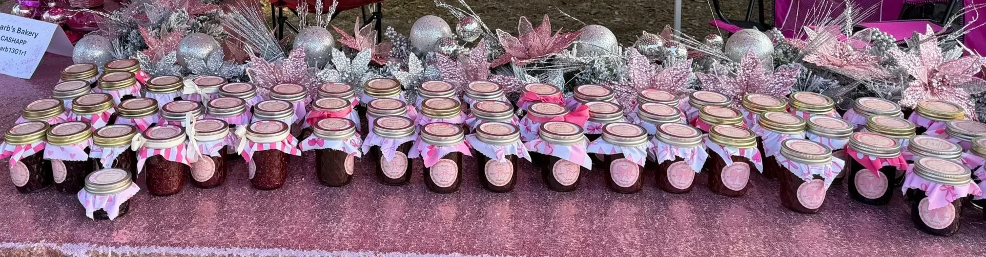 A display of small jars, possibly of jam, arranged on a pink surface with decorative Christmas-themed ornaments and pink flowers in the background.