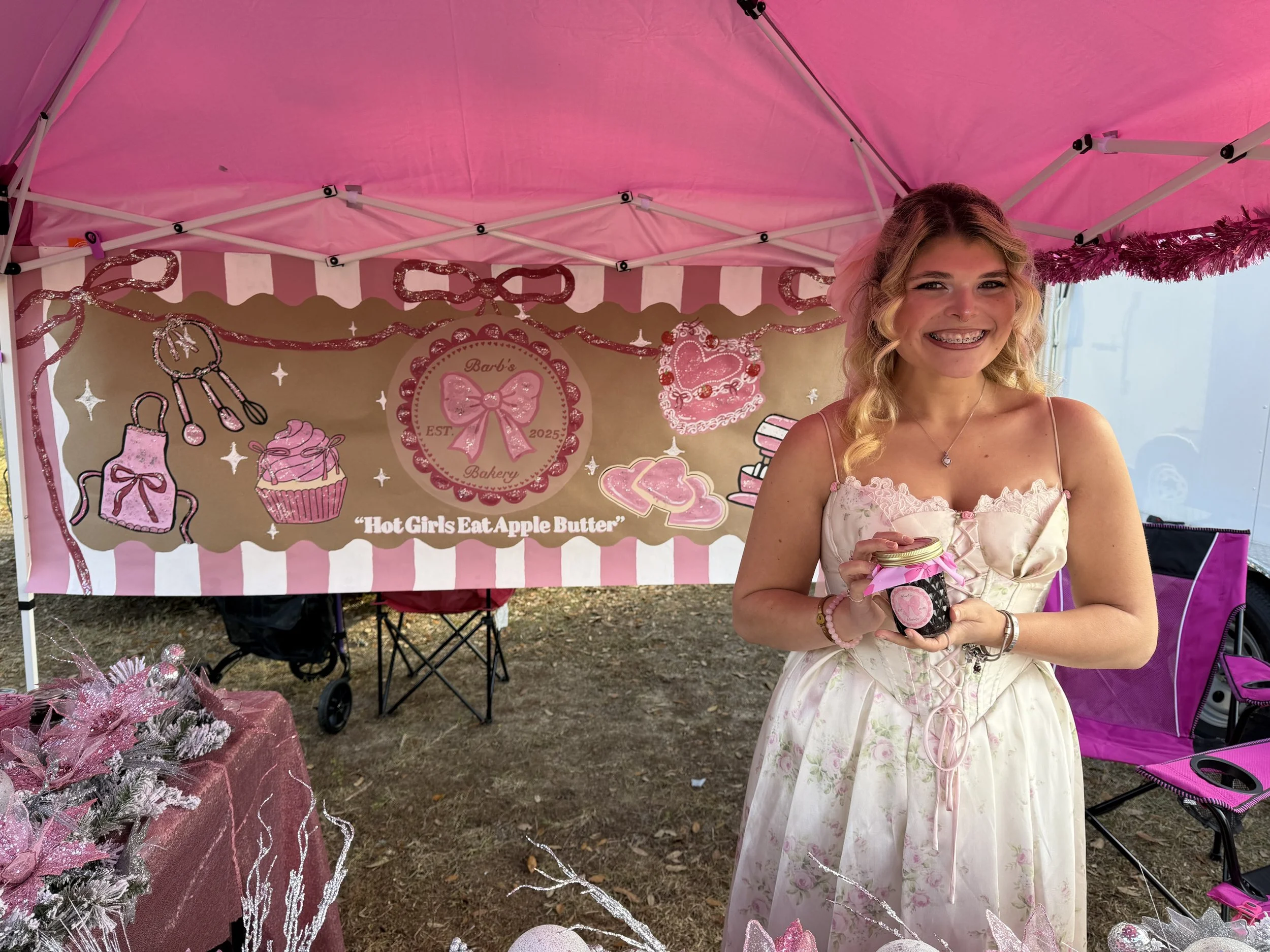 A young woman with blonde curly hair and braces wearing a white dress with pink floral accents stands in front of a bakery booth decorated in pink and white. She is holding a jar of apple butter with a pink ribbon. The booth banner reads 'Barb's Bake