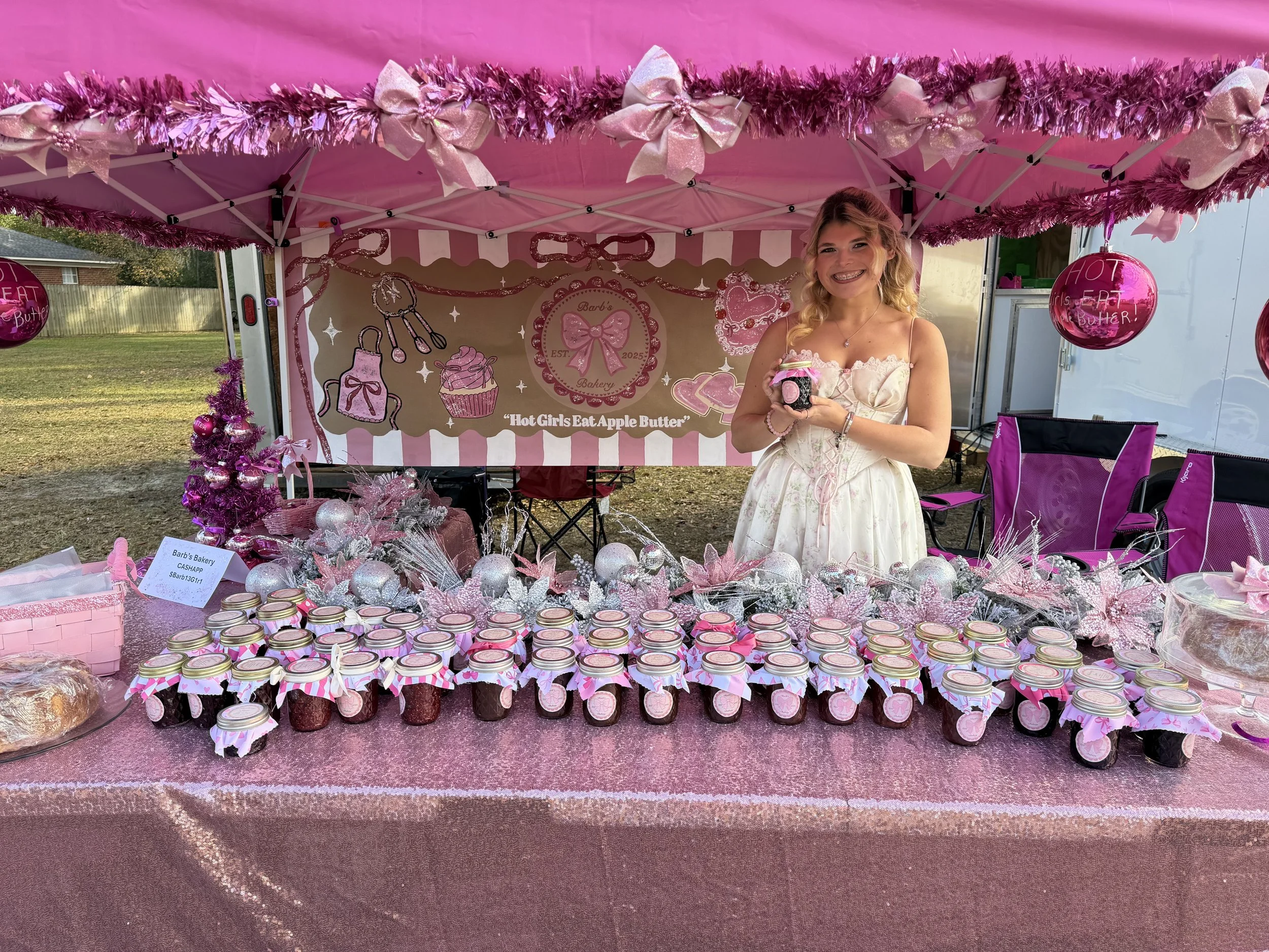 A young woman in a white dress standing behind a table with pink-themed baked goods and decorations at an outdoor bakery booth, with a pink canopy and festive decorations.