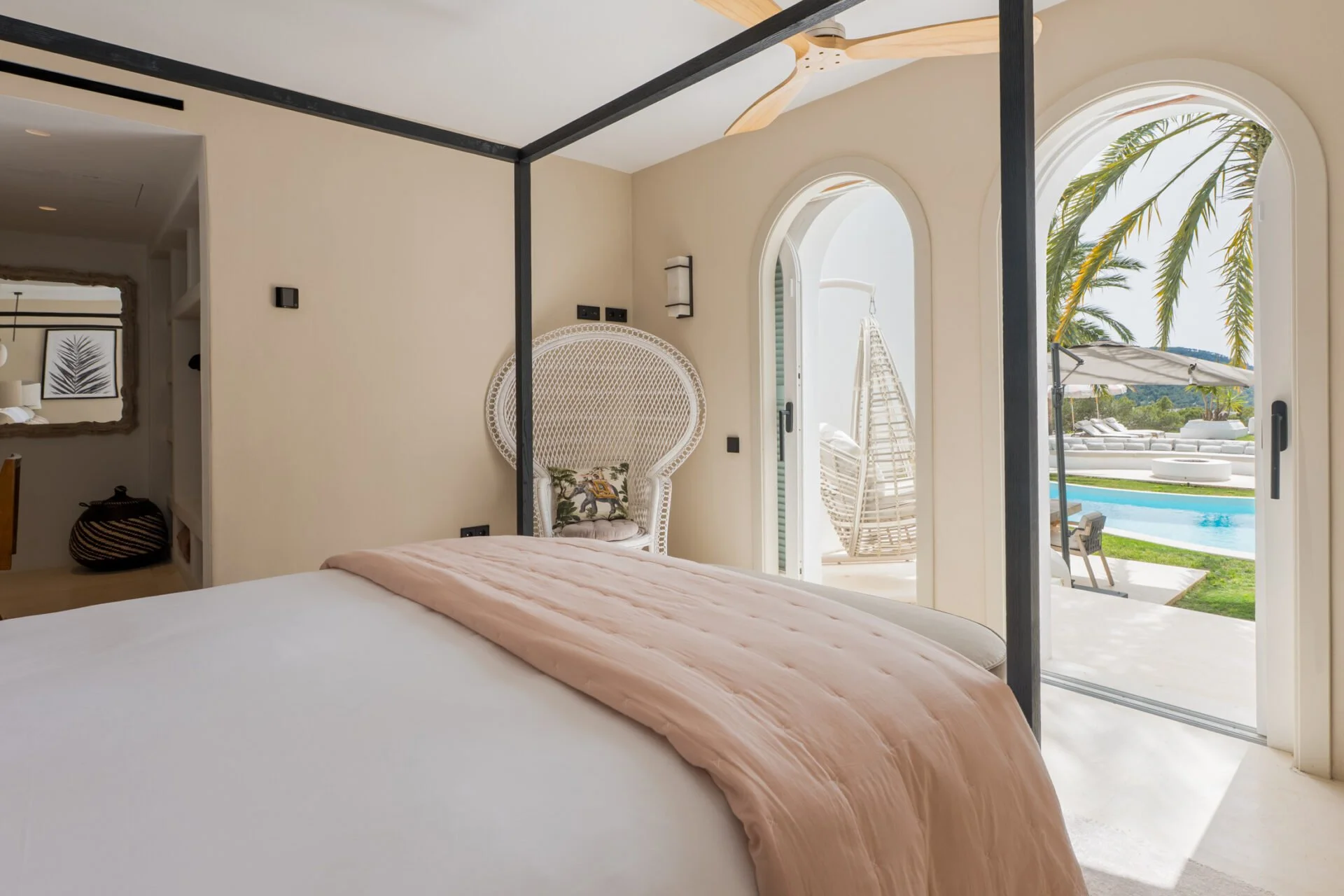 Bedroom with large arched glass door leading to patio with outdoor pool, palm trees, and lounge chairs, with a ceiling fan and decorative chair inside.