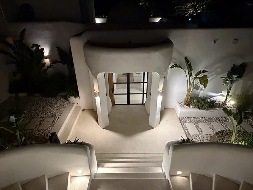 Nighttime view of a modern Ibiza villa entrance, white architectural building with stairs, plants, and decorative lighting.