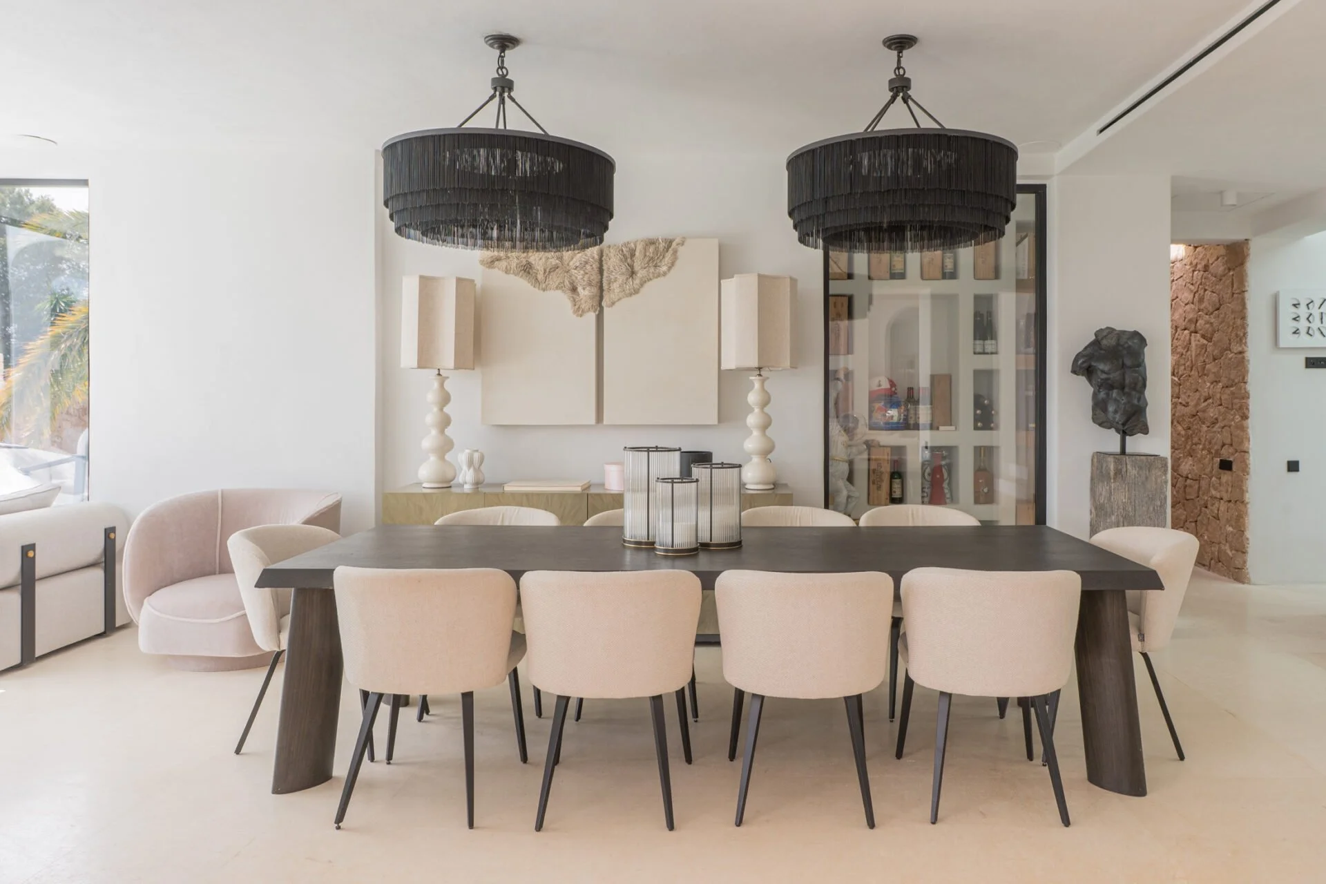 Modern dining room in Ibiza villa with a large dark wood table, surrounded by beige upholstered chairs. Two large black pendant lights hang from the ceiling. In the background, there are white and beige decor pieces, abstract artwork, a glass display