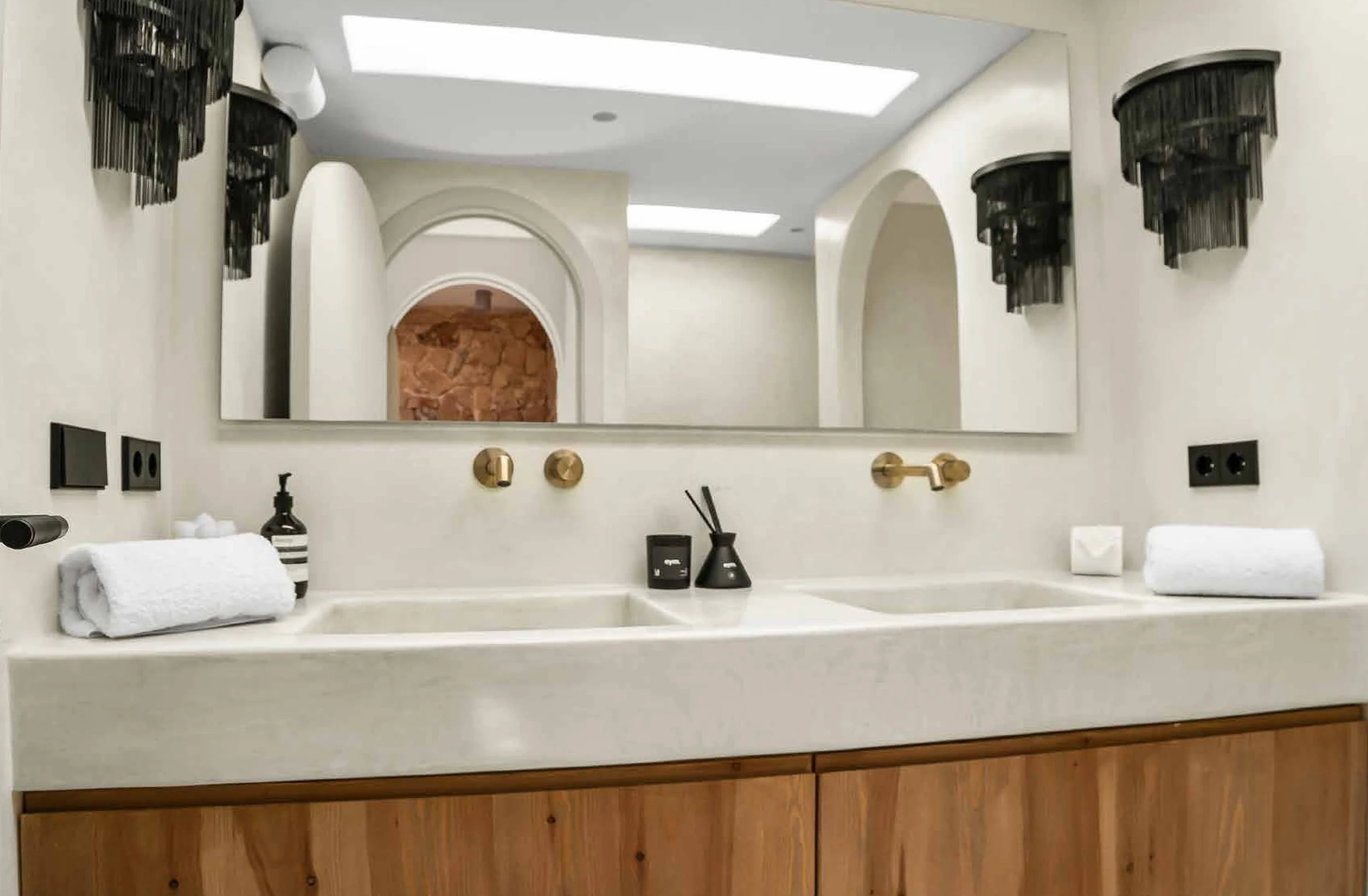 A modern bathroom vanity with two sinks, a large mirror, black and gold fixtures, black wall sconces, and decorative items including soap dispensers, towels, and reed diffusers.
