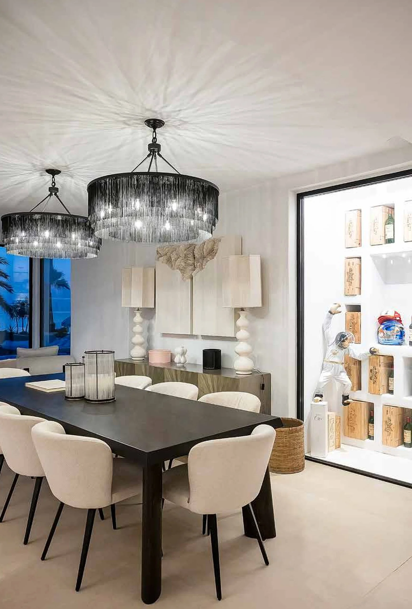Modern dining room with black table, beige upholstered chairs, two black chandeliers with fringe, white wall with decorative lamps, and a shelving unit with vintage boxes, figurines, and bottles.