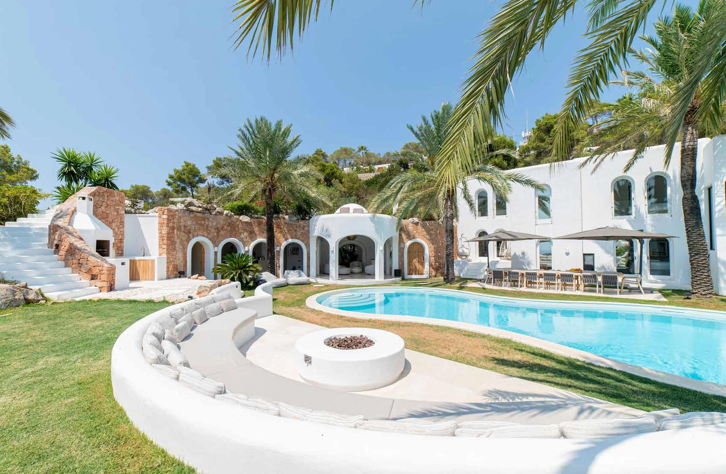 Luxury outdoor residential area with a swimming pool, surrounded by white and stone buildings, palm trees, and outdoor seating under umbrellas