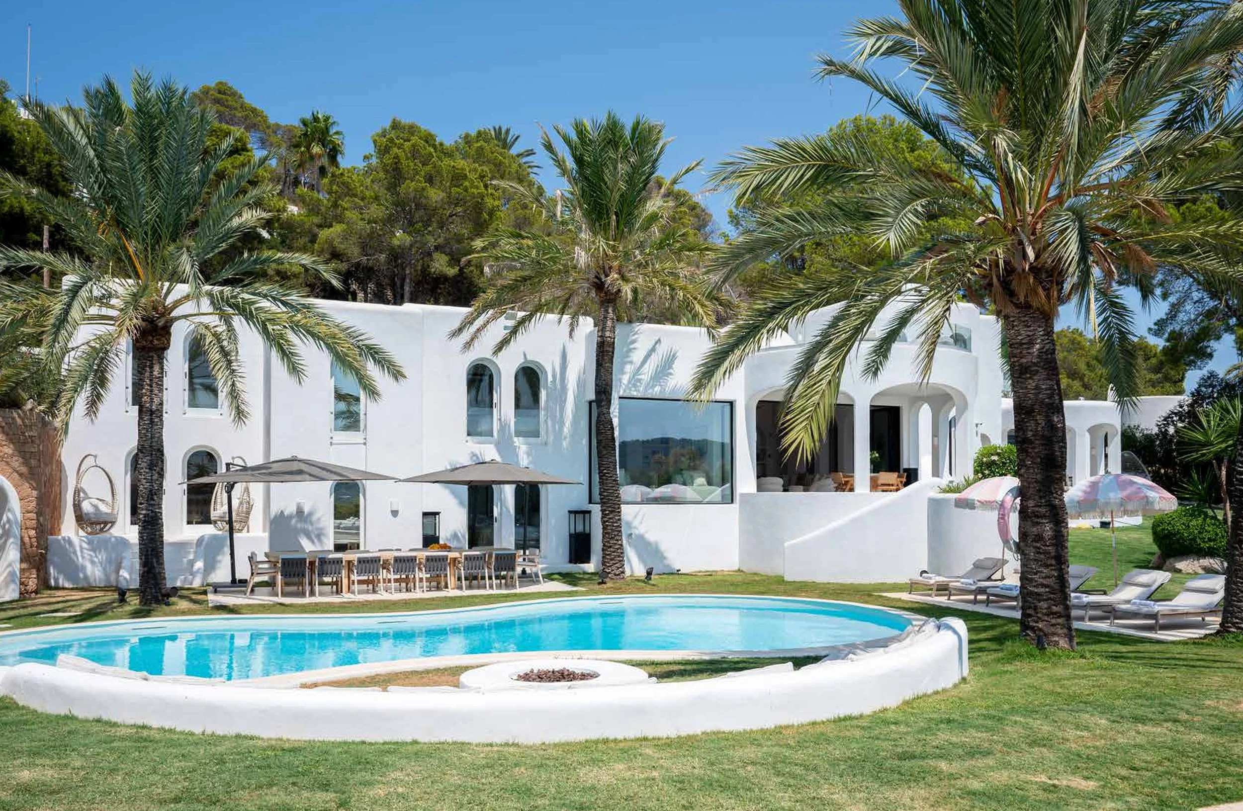Luxury white villa with palm trees, outdoor pool, lounge chairs, umbrellas, and patio furniture under a clear blue sky.