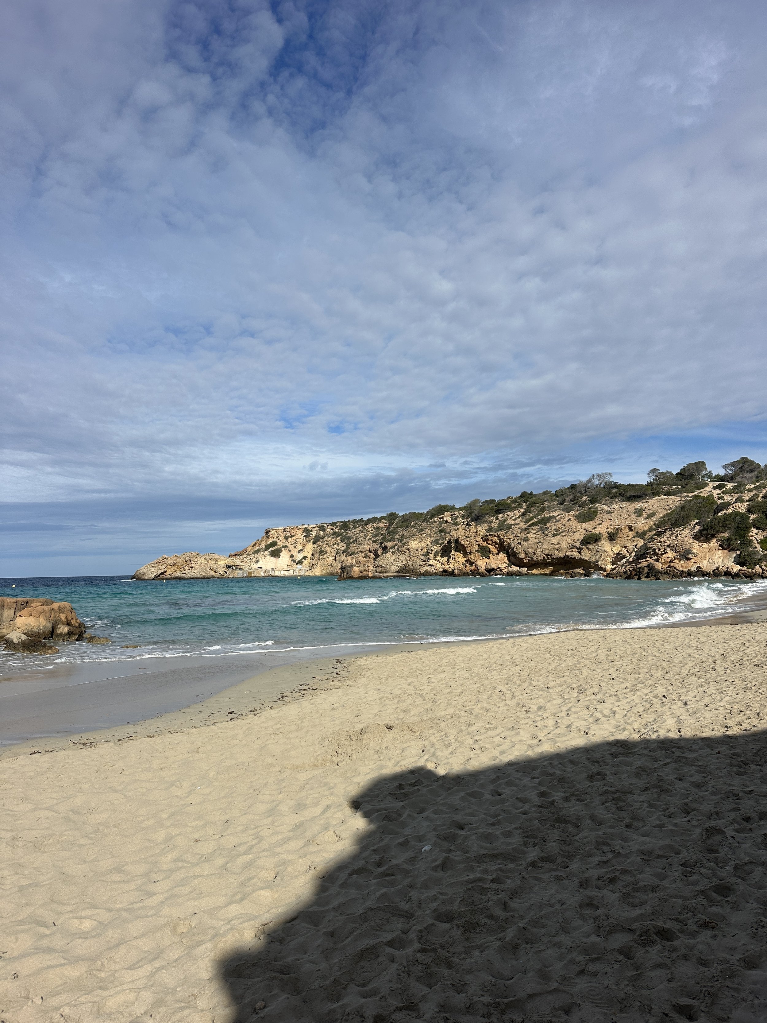 Sunny beach with sandy shore, turquoise water, rocky cliffs, and partly cloudy sky.