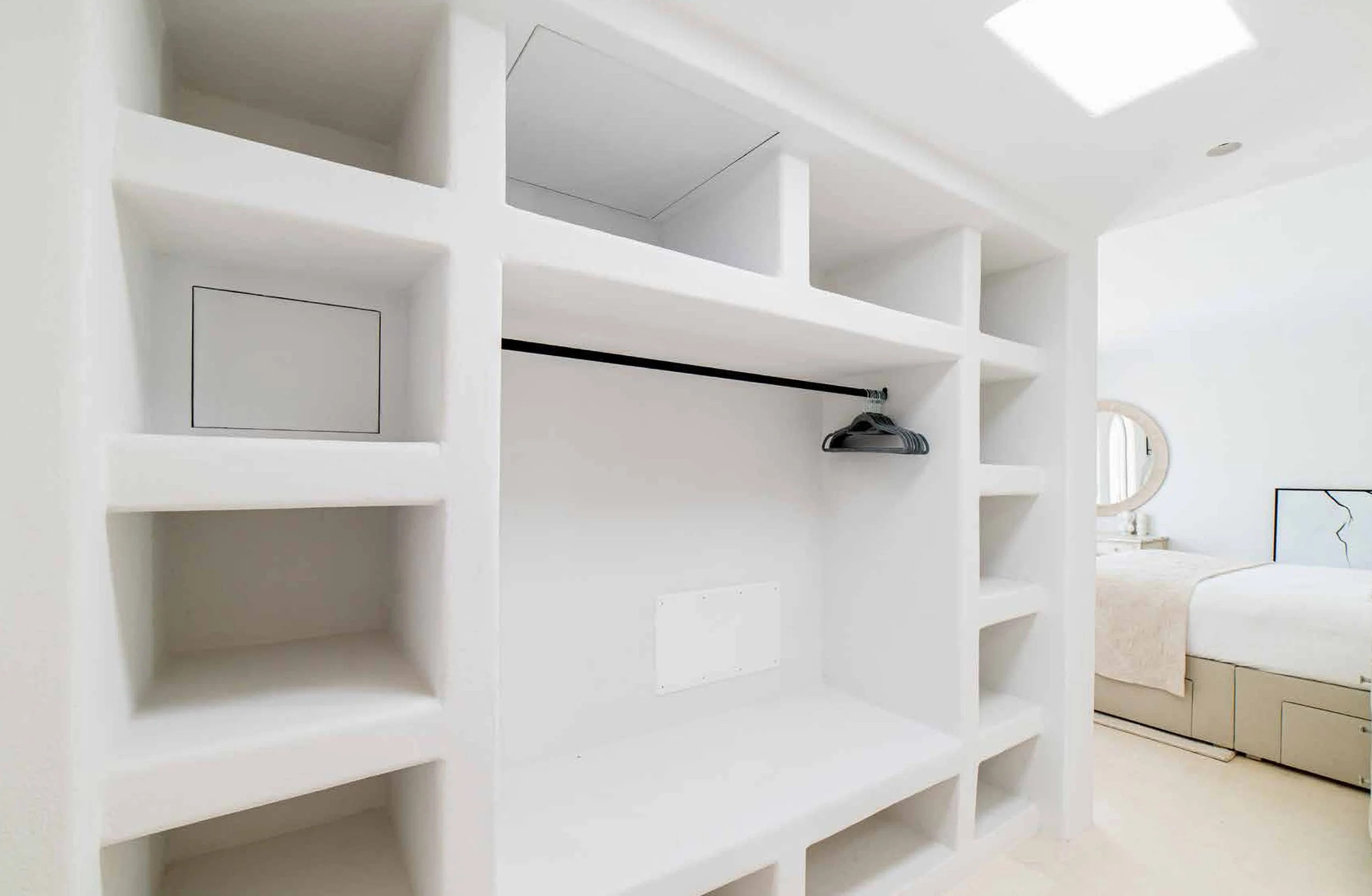 Empty built-in white shelving unit in a bright bedroom, with a few empty sections and a hanging rod. In the background, a bed with a beige headboard and a round mirror on a dresser are visible.