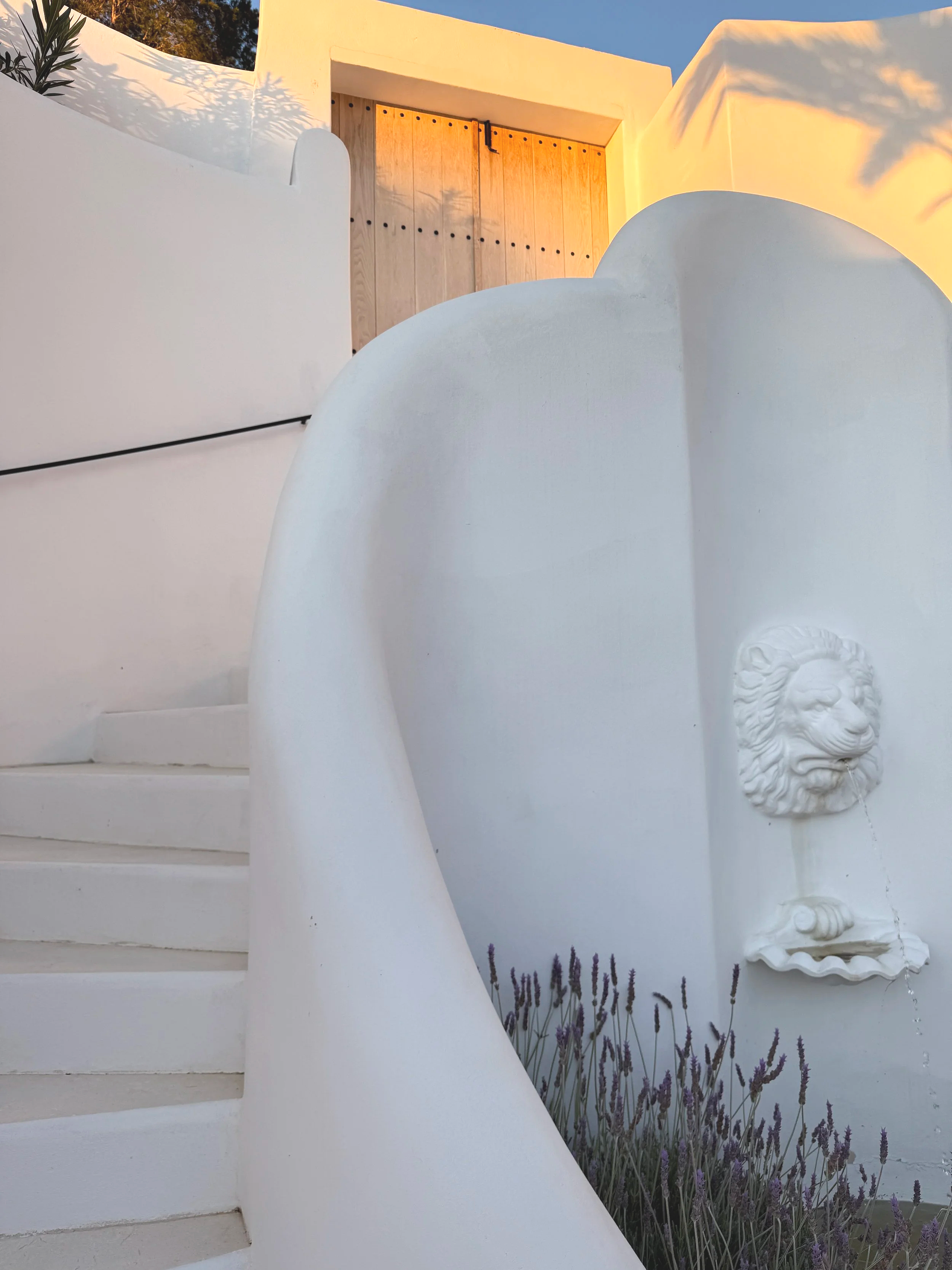 White stucco building with a curved stairway, a lion head water fountain, purple lavender plants, and a wooden door on a second floor.